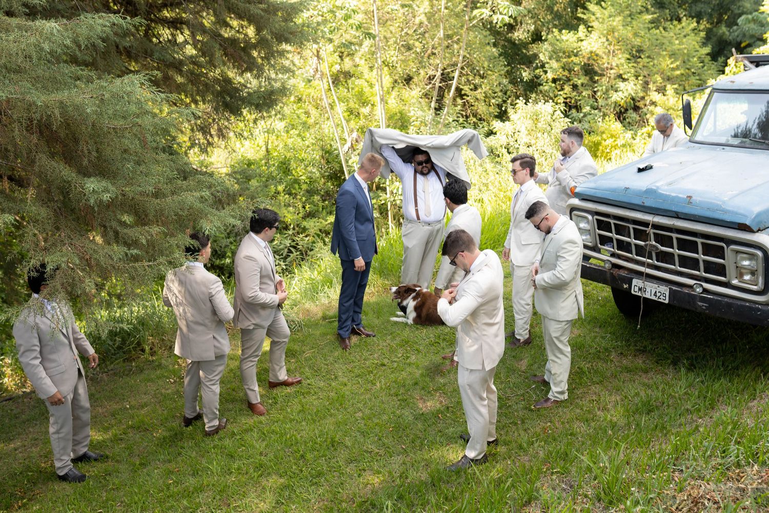 Groomsmen in tan suits gather outdoors near a blue vintage truck surrounded by lush green trees before a wedding.