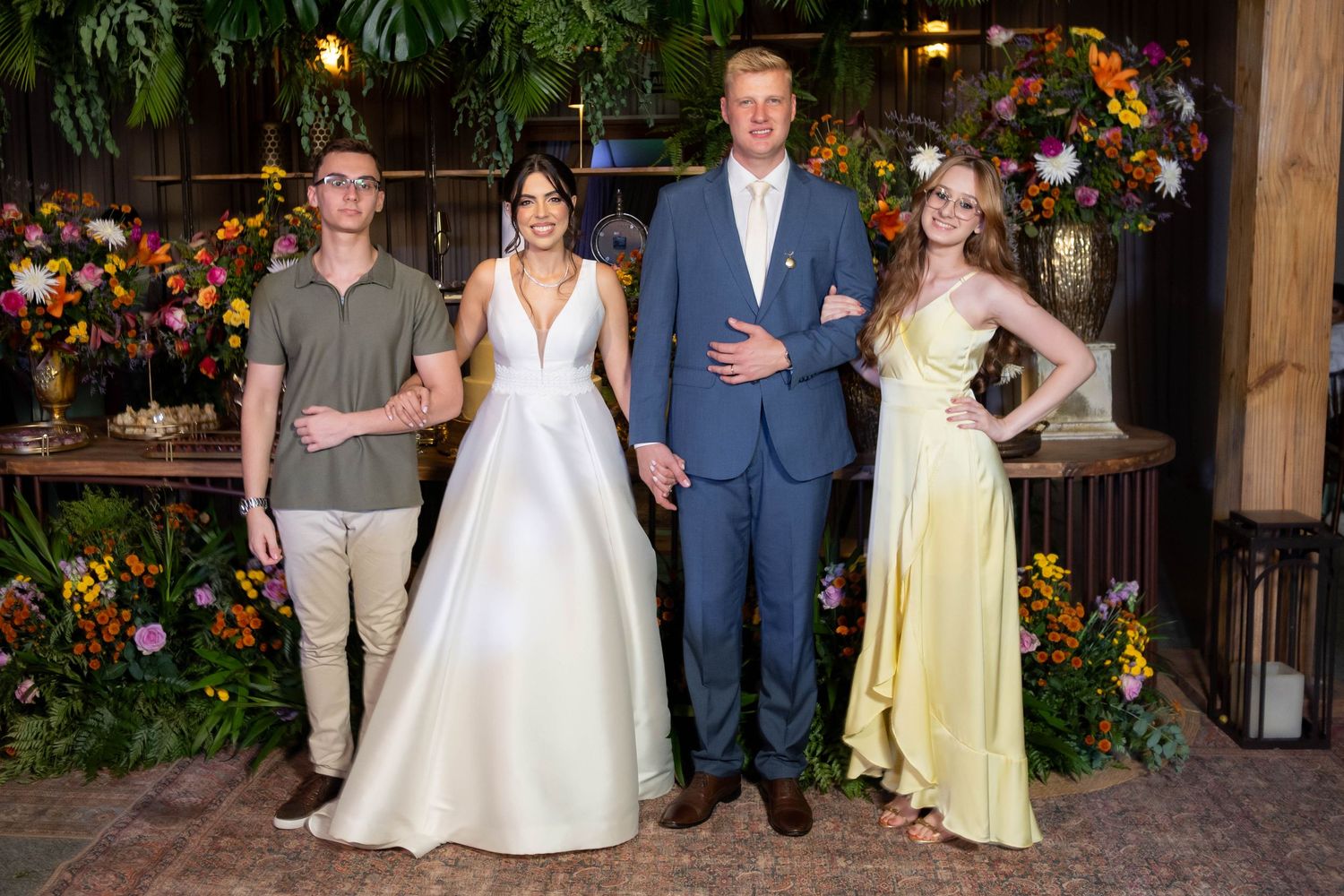 A bride in a white gown and groom in blue suit pose with two guests amid floral decor at a wedding reception.
