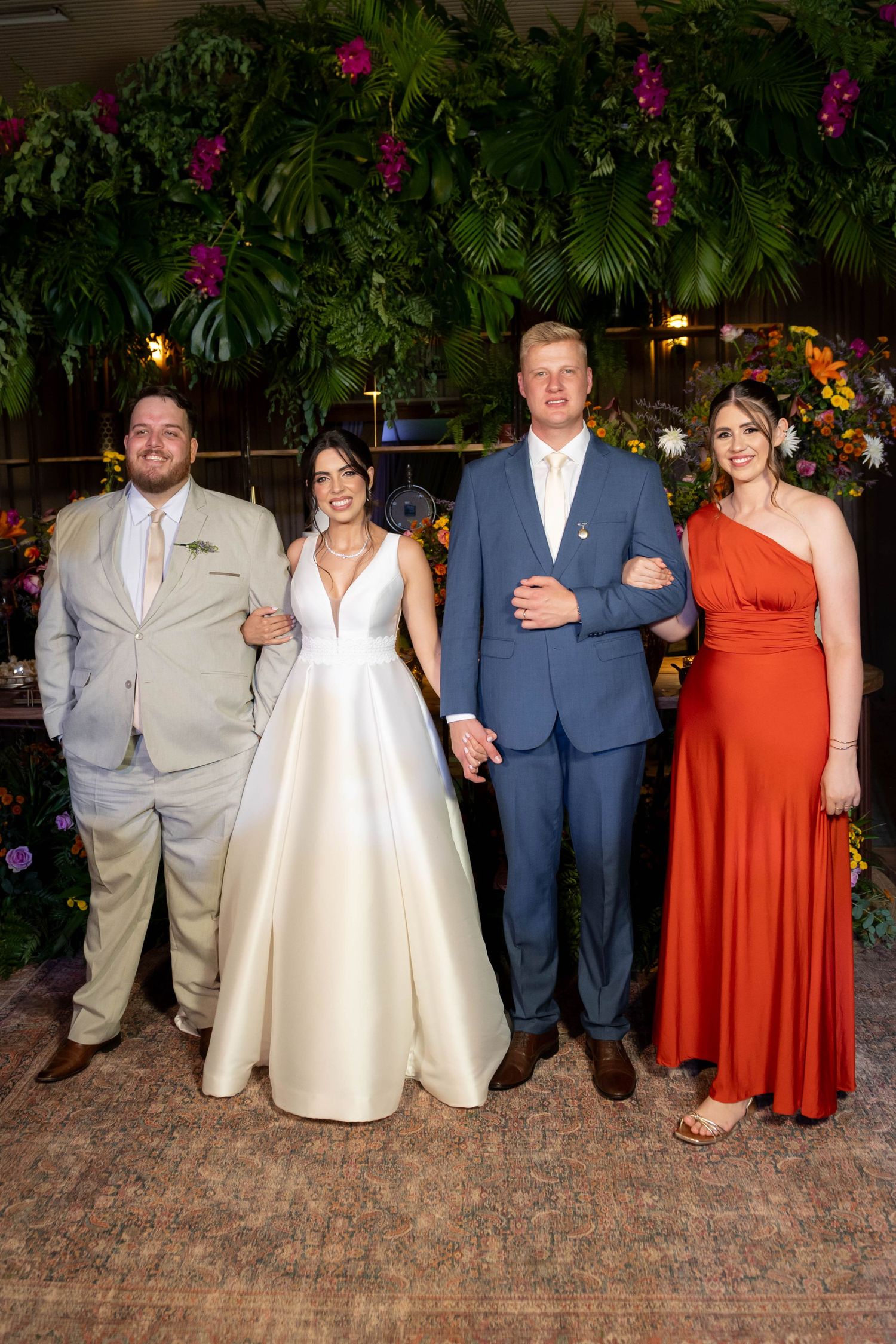 Two couples pose together at a wedding, bride in white gown, groom in blue suit, guests in gray suit and red dress.
