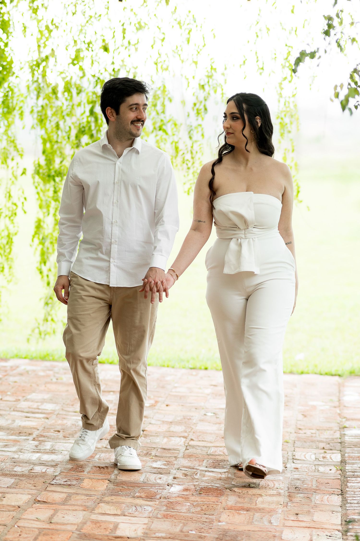 Couple in white outfits walking hand-in-hand on brick path under green weeping willow tree.
