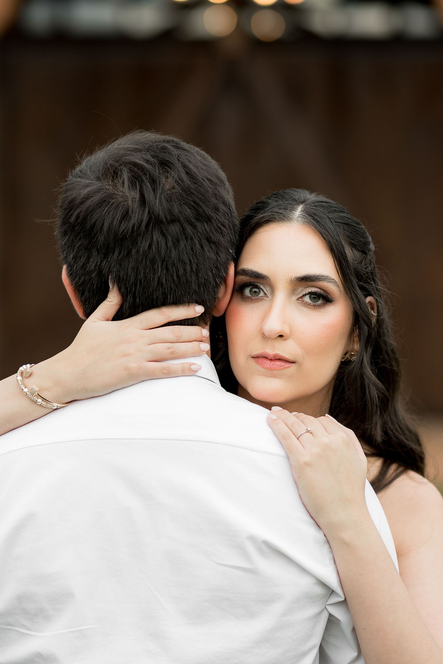 Bride with dark wavy hair embracing groom in white shirt during romantic outdoor portrait.
