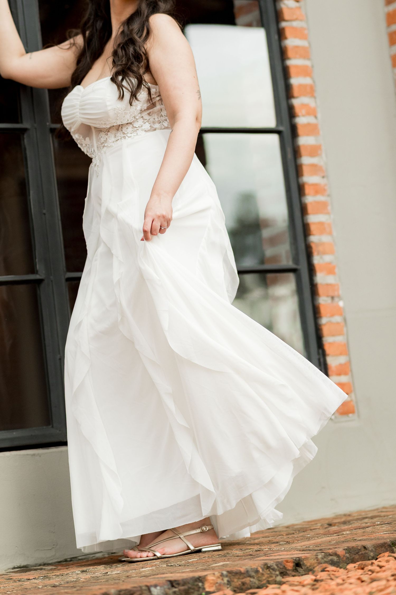 Bride in a strapless white wedding gown with ruffled skirt and beaded bodice posing outdoors near brick wall.