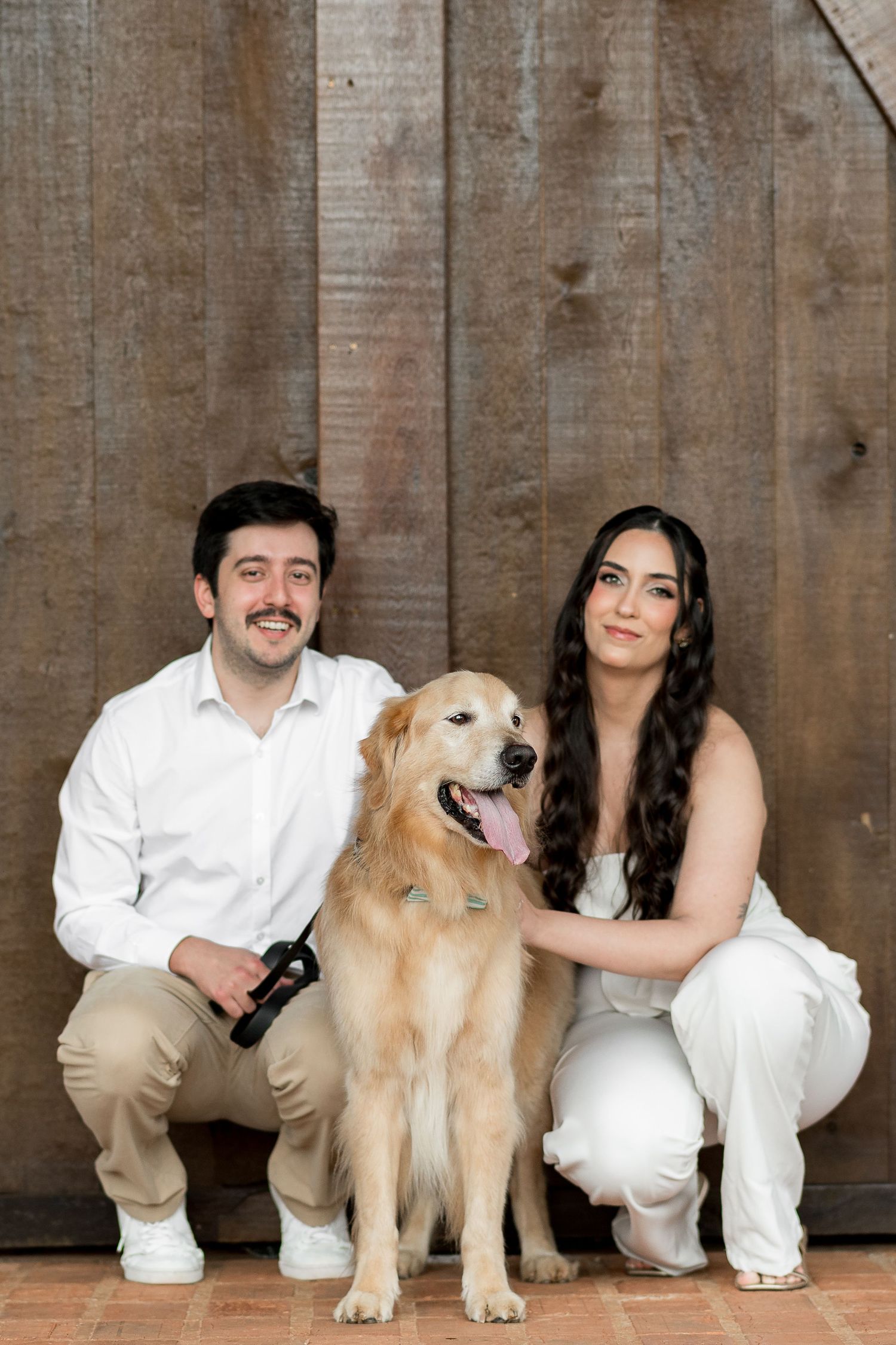 Couple in white outfits posing with a golden retriever dog against a rustic wooden wall backdrop.