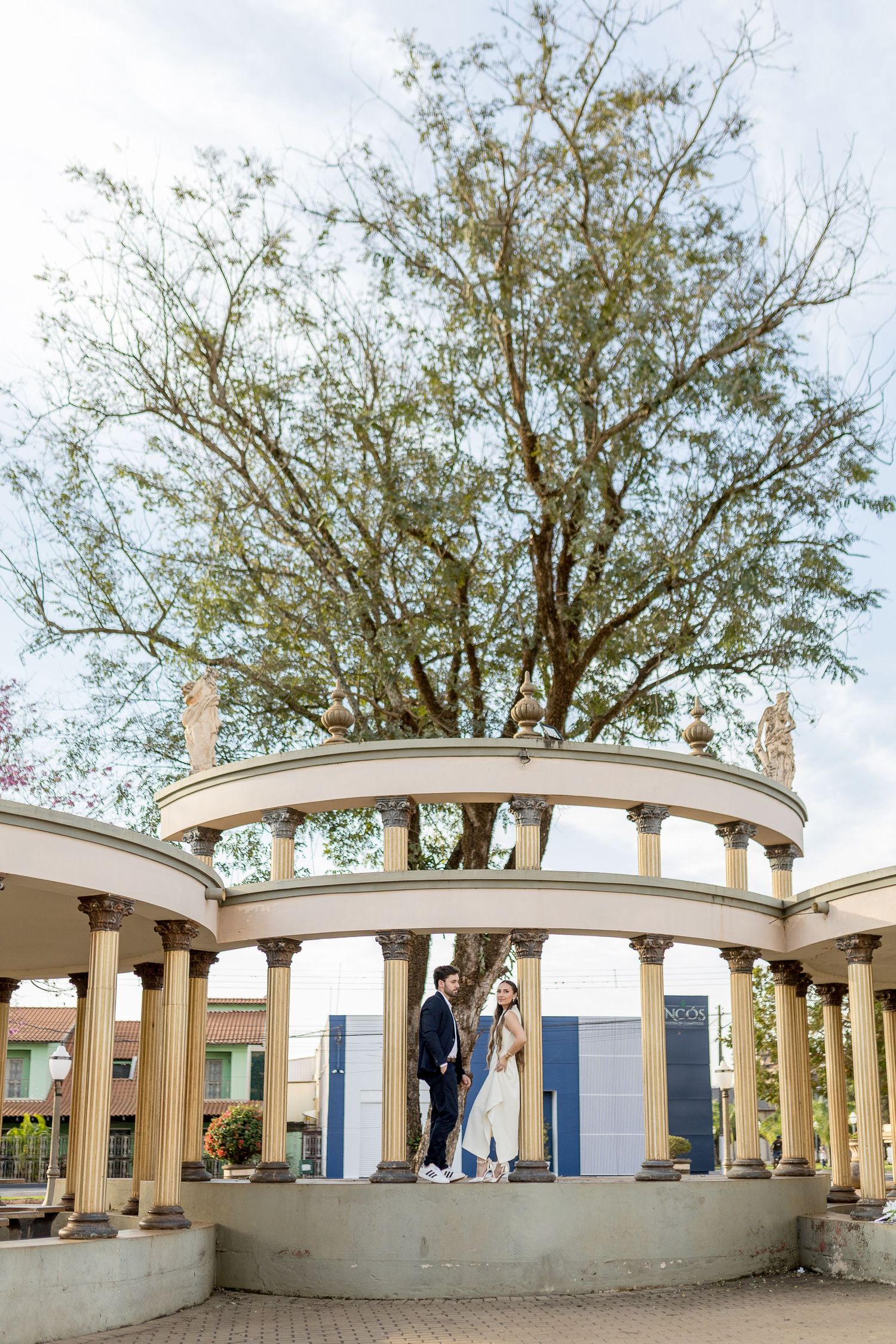 Couple standing together in a classical colonnade with tall columns and a large tree backdrop at an outdoor venue.