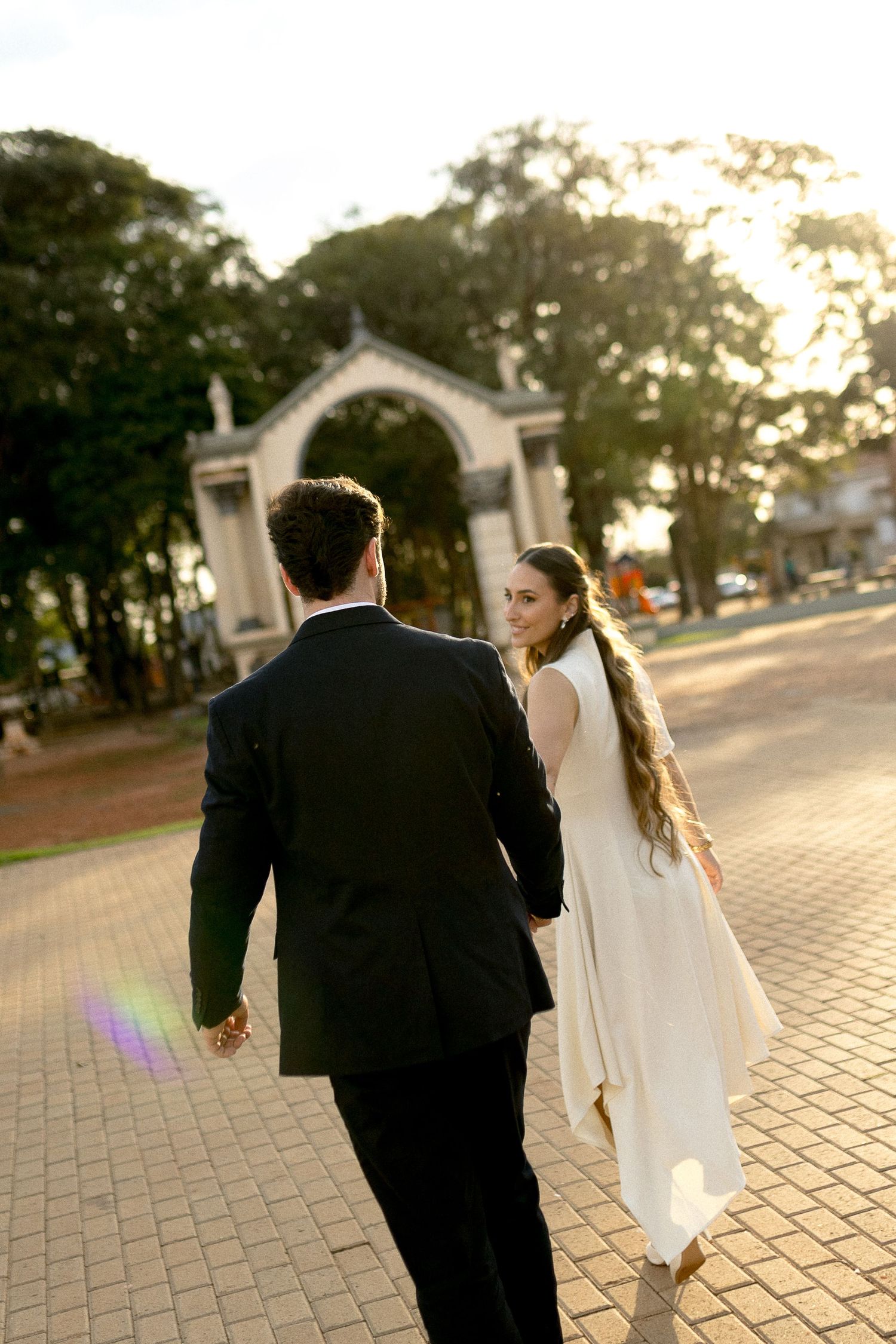 A bride and groom walk hand-in-hand toward an ornate arch in a sunlit park, as she glances back with a smile.
