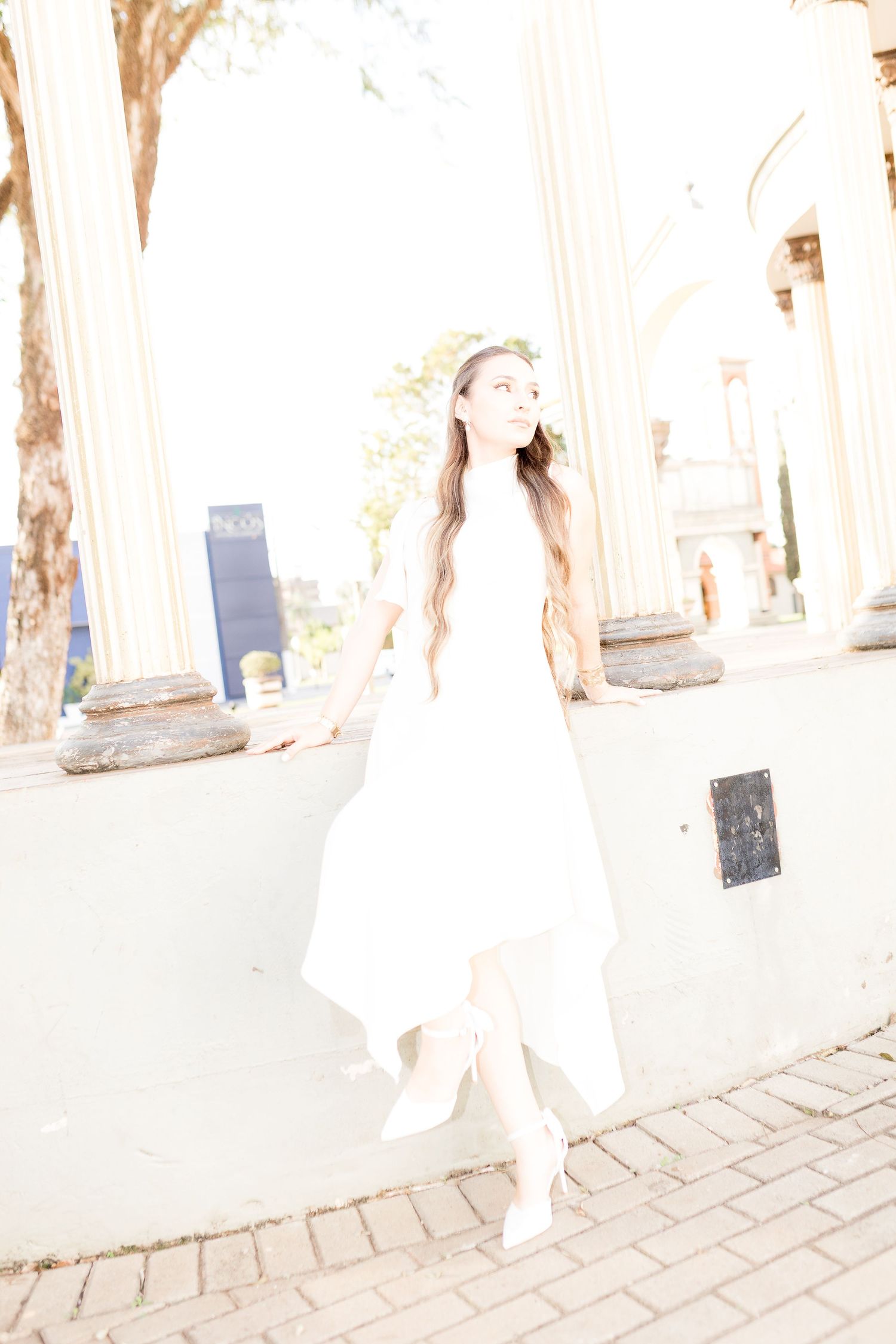 Woman in white dress posing near classical columns in bright sunlight.