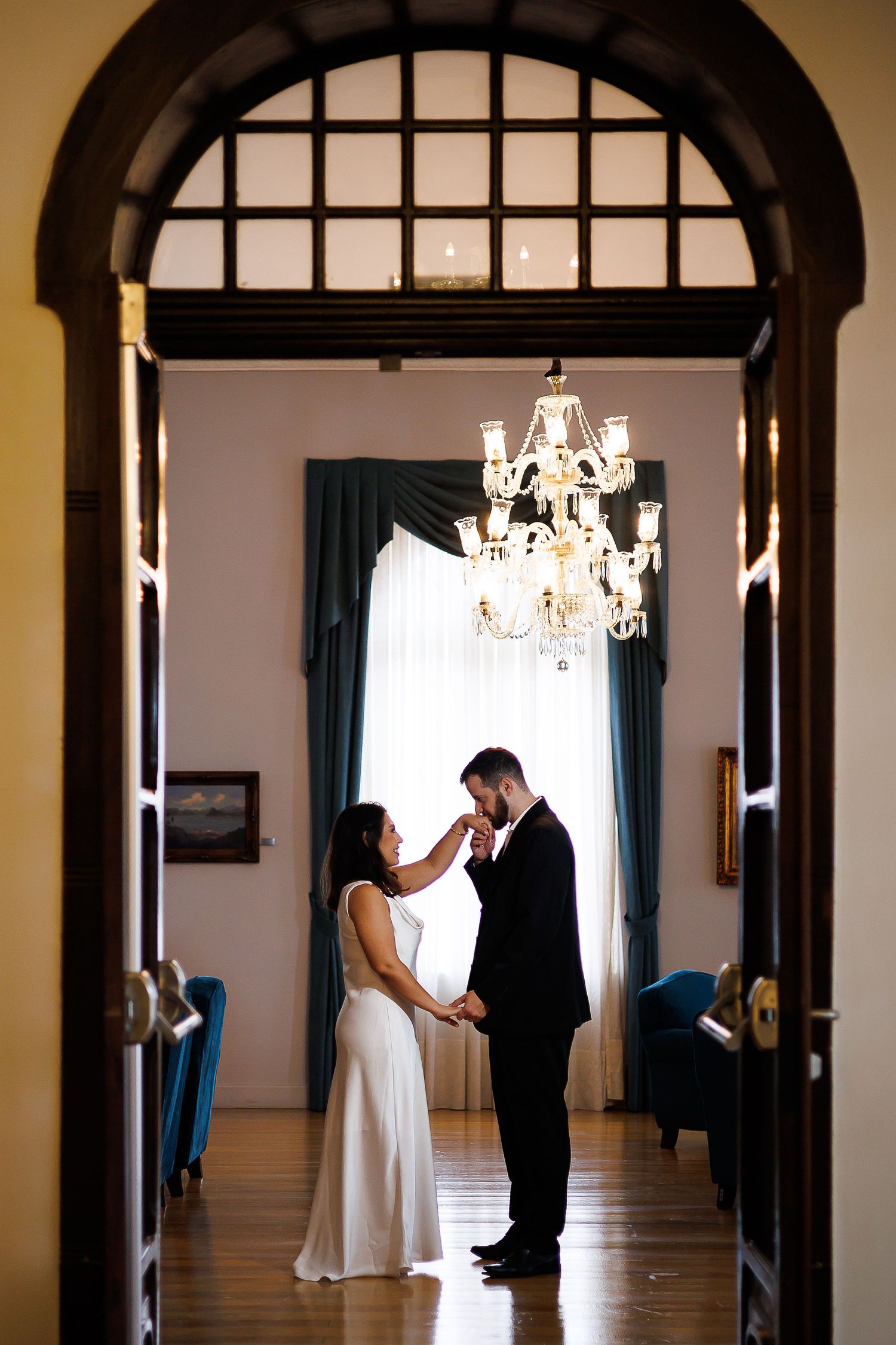 Bride and groom share a romantic moment under a crystal chandelier in an elegant room with arched doorway.