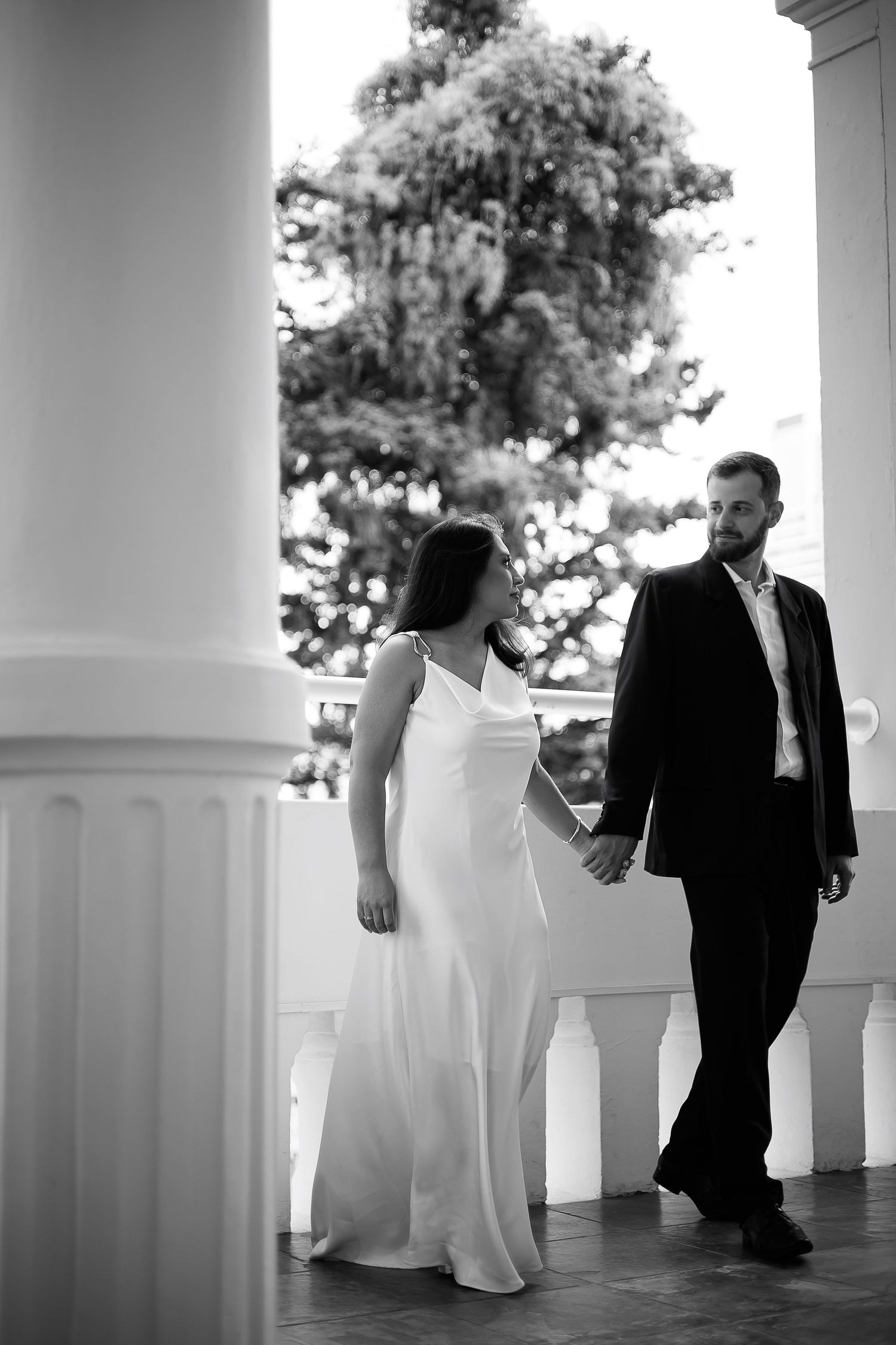 Black and white photo of a bride in white gown and groom in suit holding hands on a elegant columned porch.