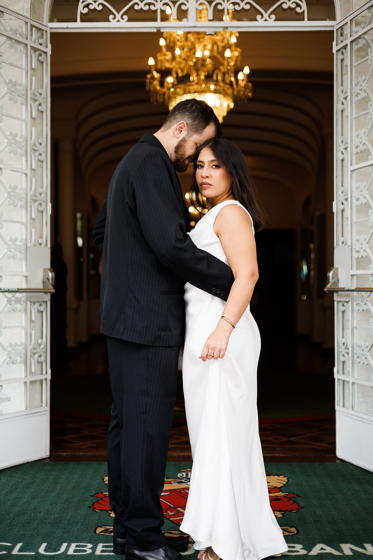 Elegant couple in formal attire embracing in a grand hall with chandelier, man in black suit, woman in white gown.