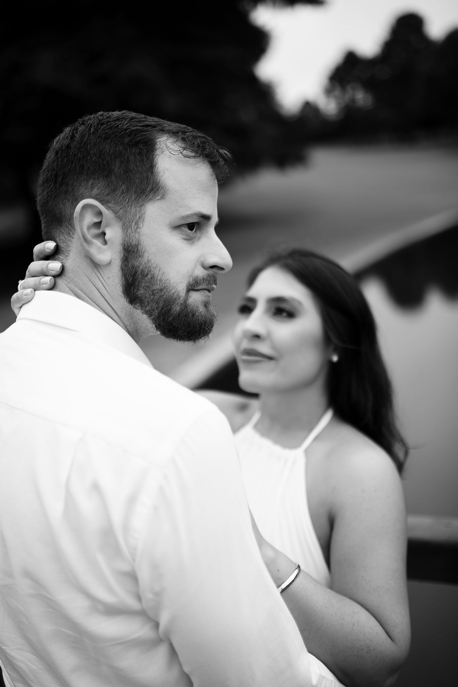Black and white photo of a bearded man and dark-haired woman embracing outdoors near a lake.