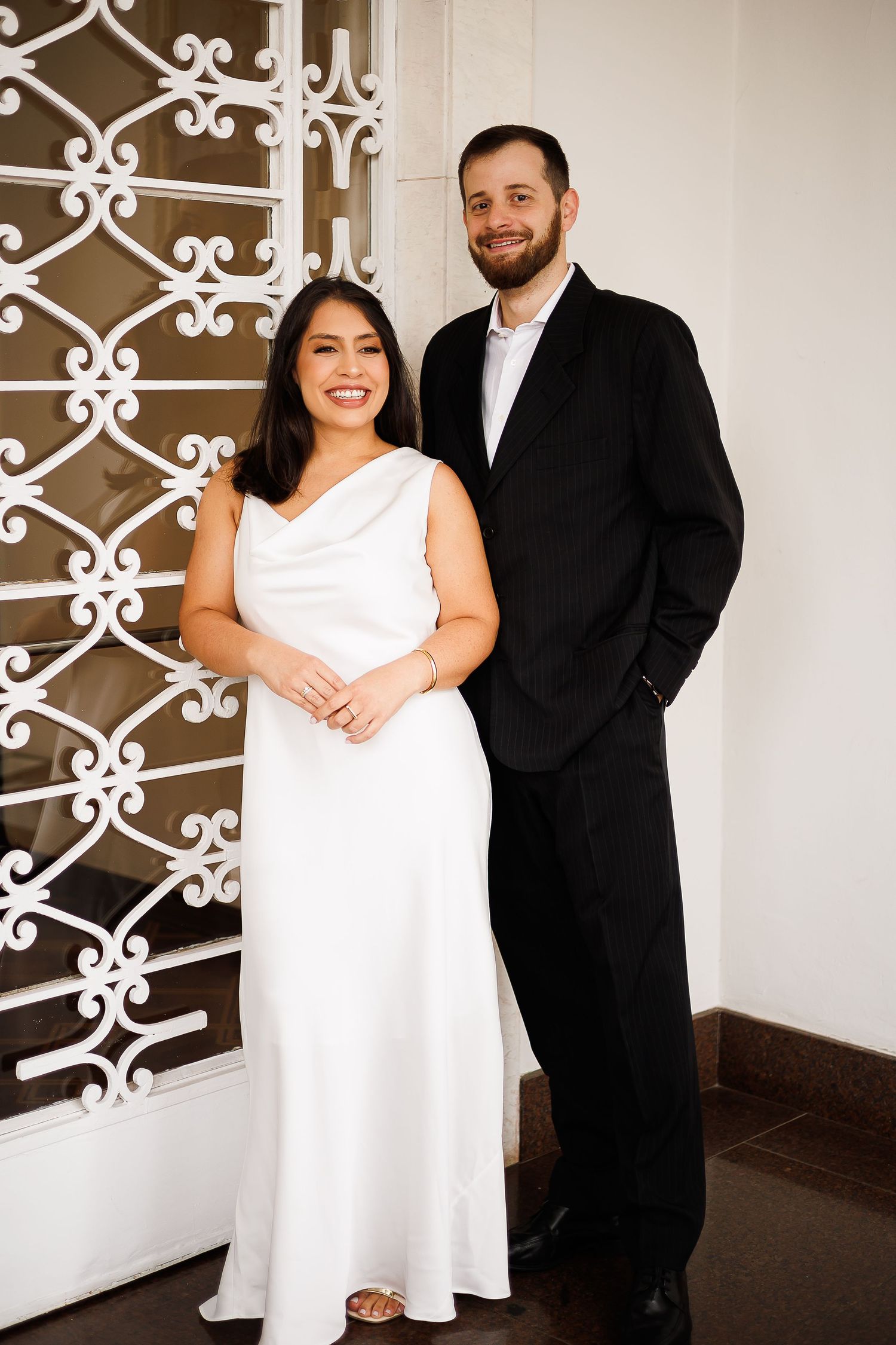 A couple poses elegantly, woman in white gown and man in black suit, smiling before ornate white ironwork door.