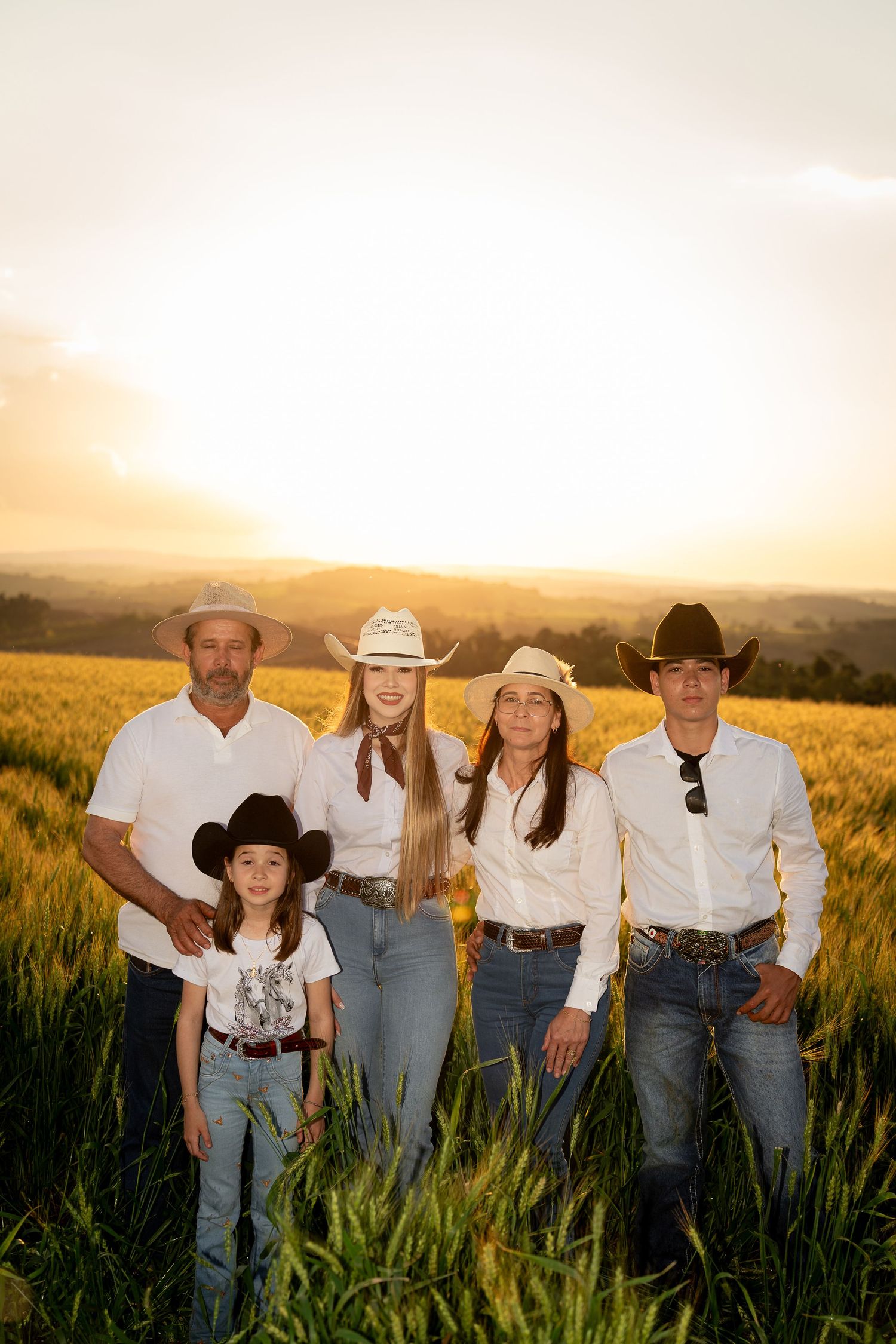 Family of five in white shirts and cowboy hats posing in a golden field at sunset with mountains in the background.