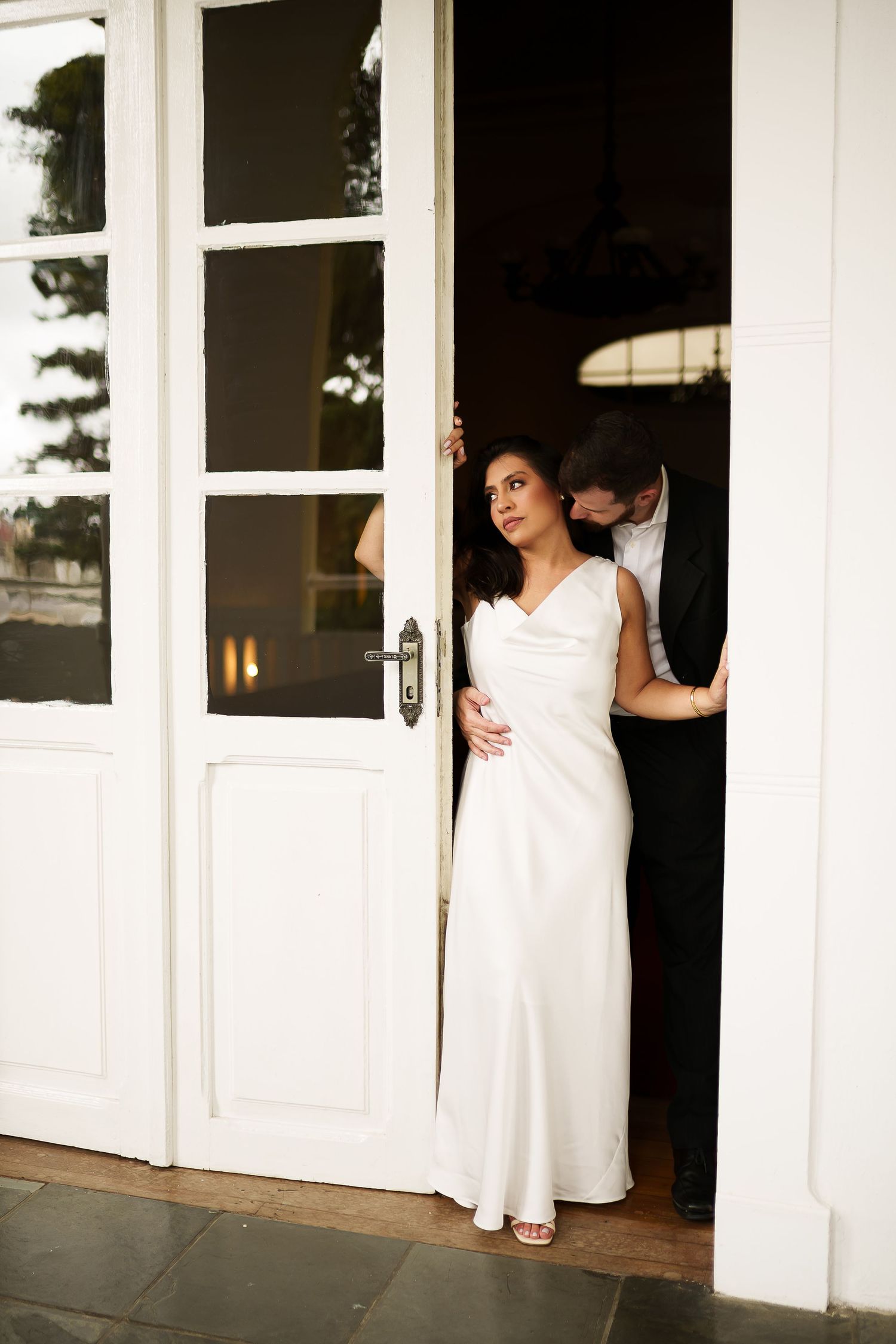 Bride in elegant white gown standing in French doors, groom in black suit behind her at a wedding venue.