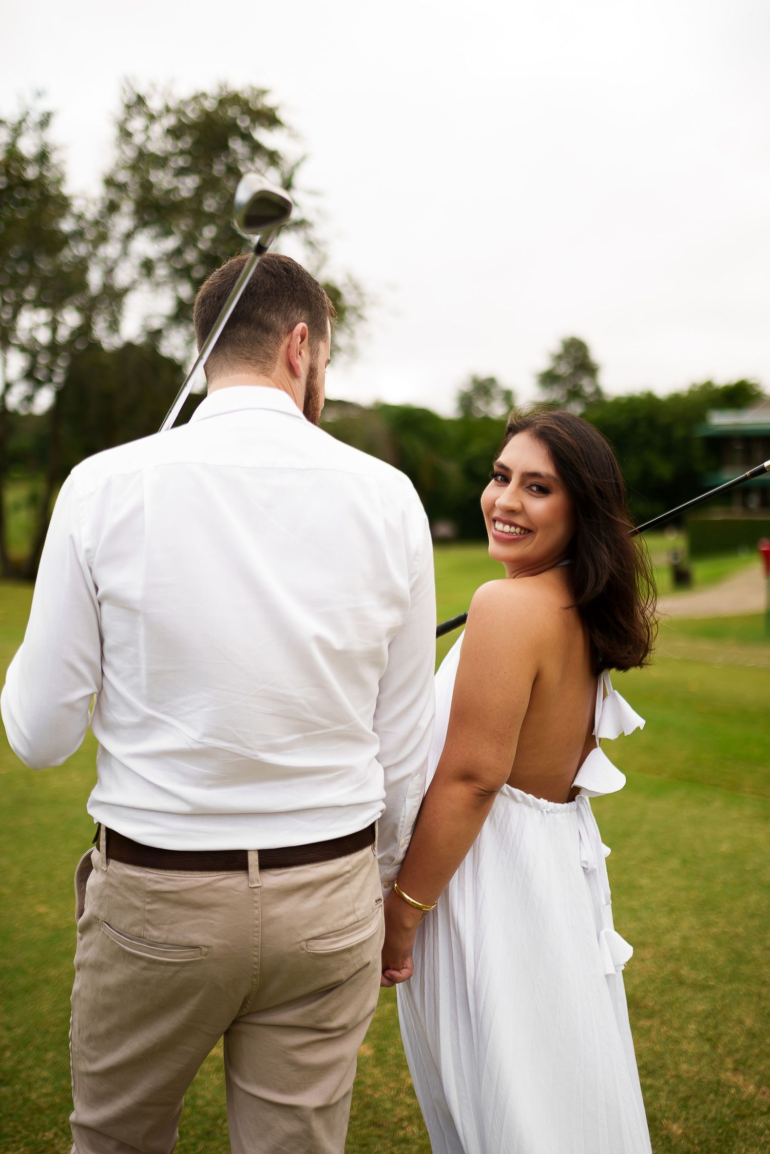 A smiling woman in a white backless dress holds hands with a man in white shirt on a golf course.