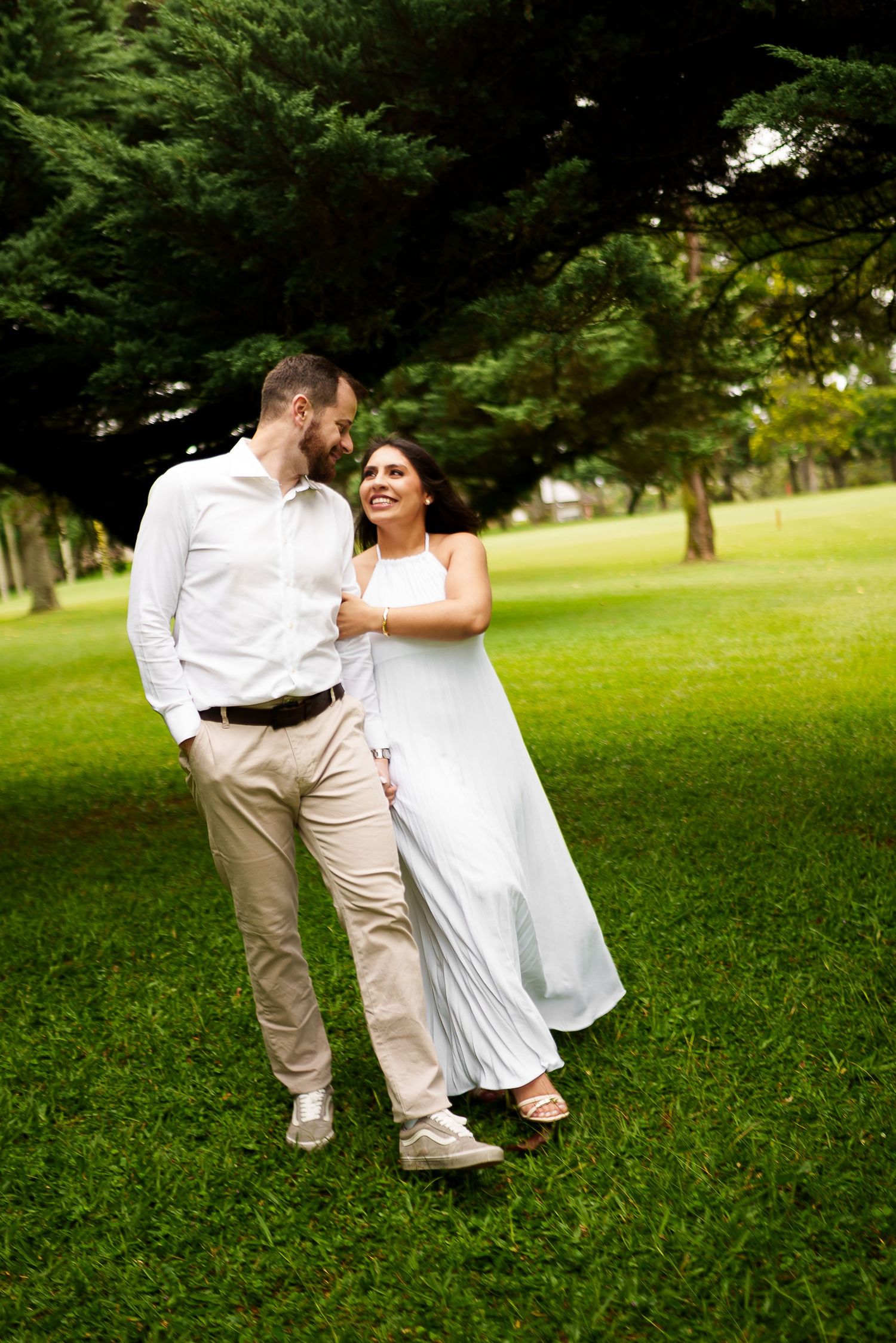 Couple in white outfits walking and laughing together on lush green grass in a park with tall trees.