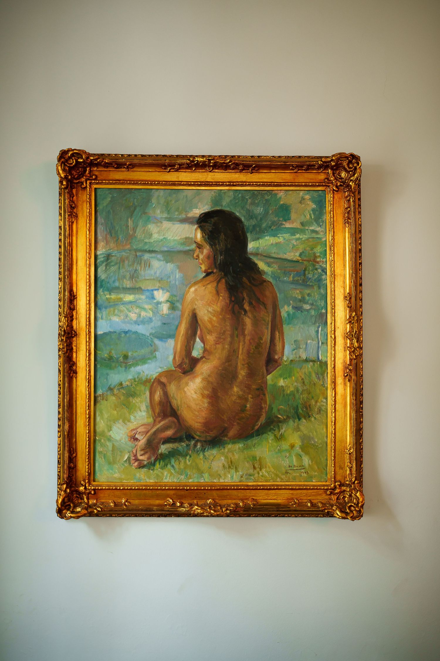 Oil painting in ornate gold frame depicting a nude figure seated outdoors near water, viewed from behind.
