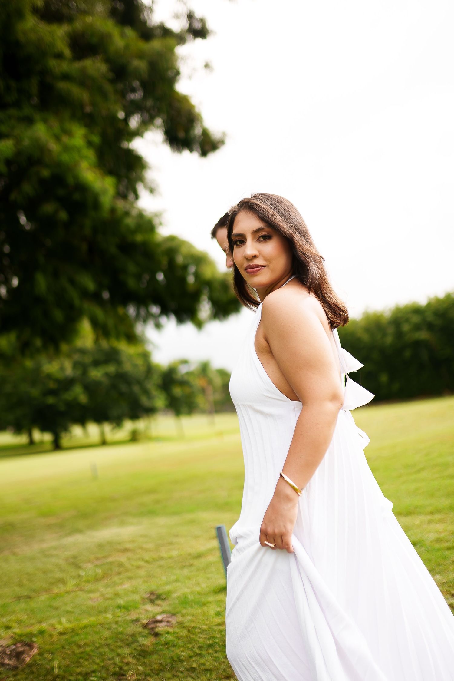 Woman in white pleated dress posing outdoors in lush green park.
