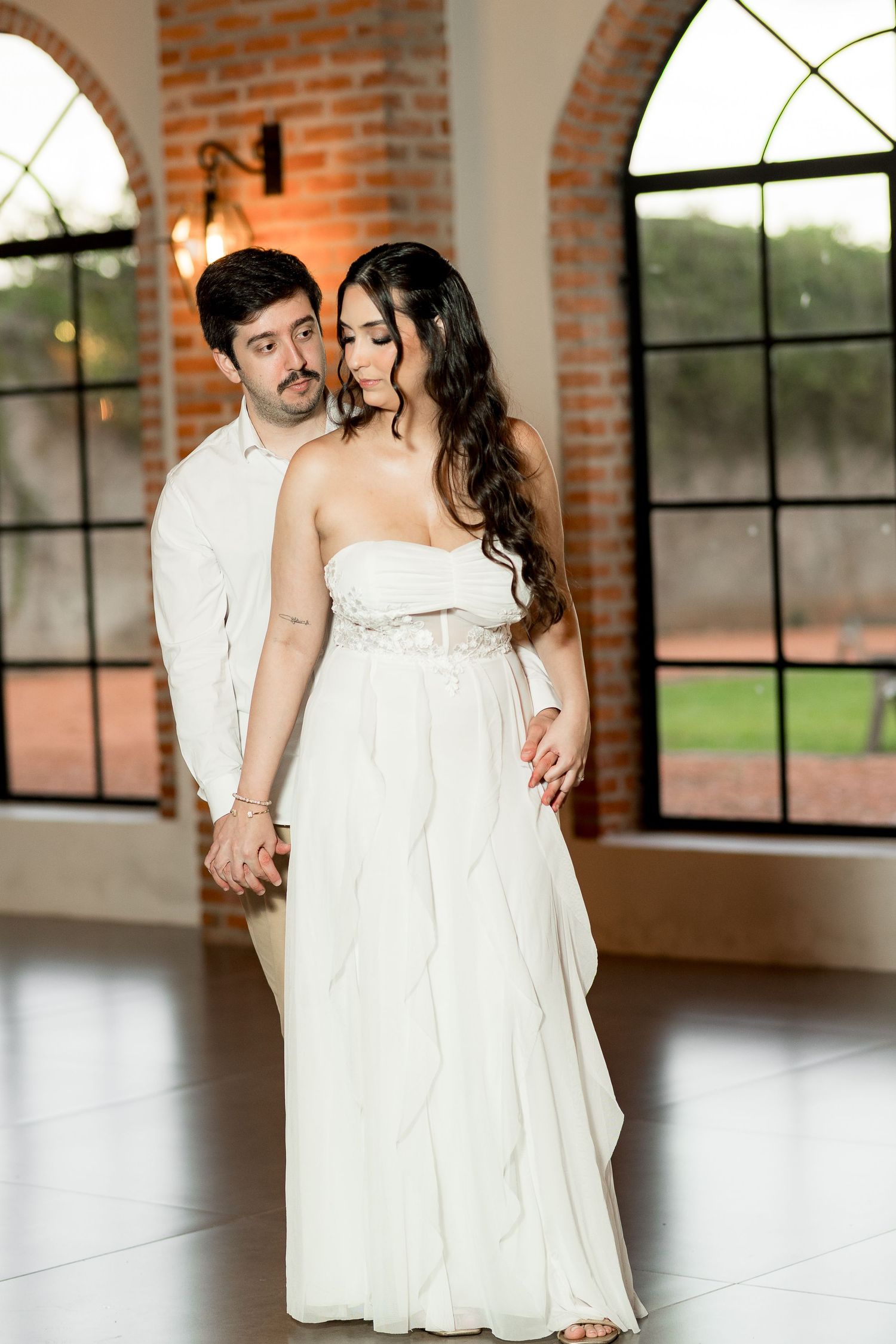 Elegant couple in white attire sharing a romantic moment in a bright venue with arched windows and brick walls.