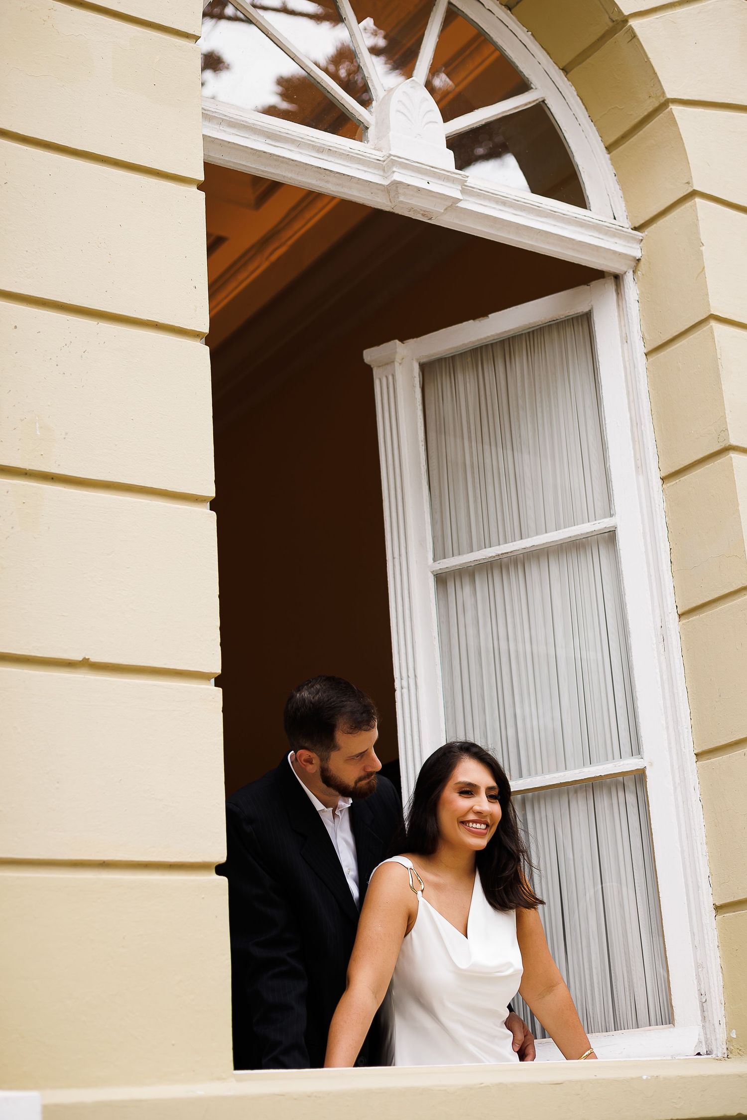Couple in wedding attire standing on a classic building balcony, bride in white dress smiling warmly.