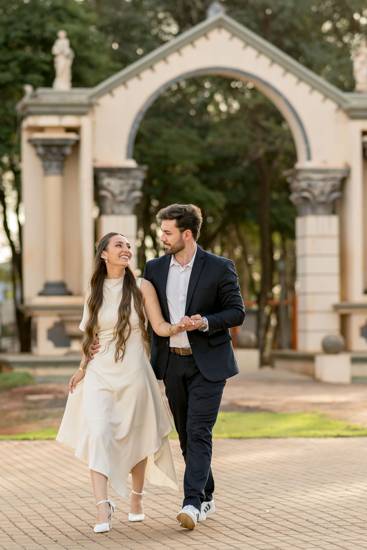 Elegant couple walking hand in hand near ornate arch, woman in white dress, man in dark suit, lush garden backdrop.
