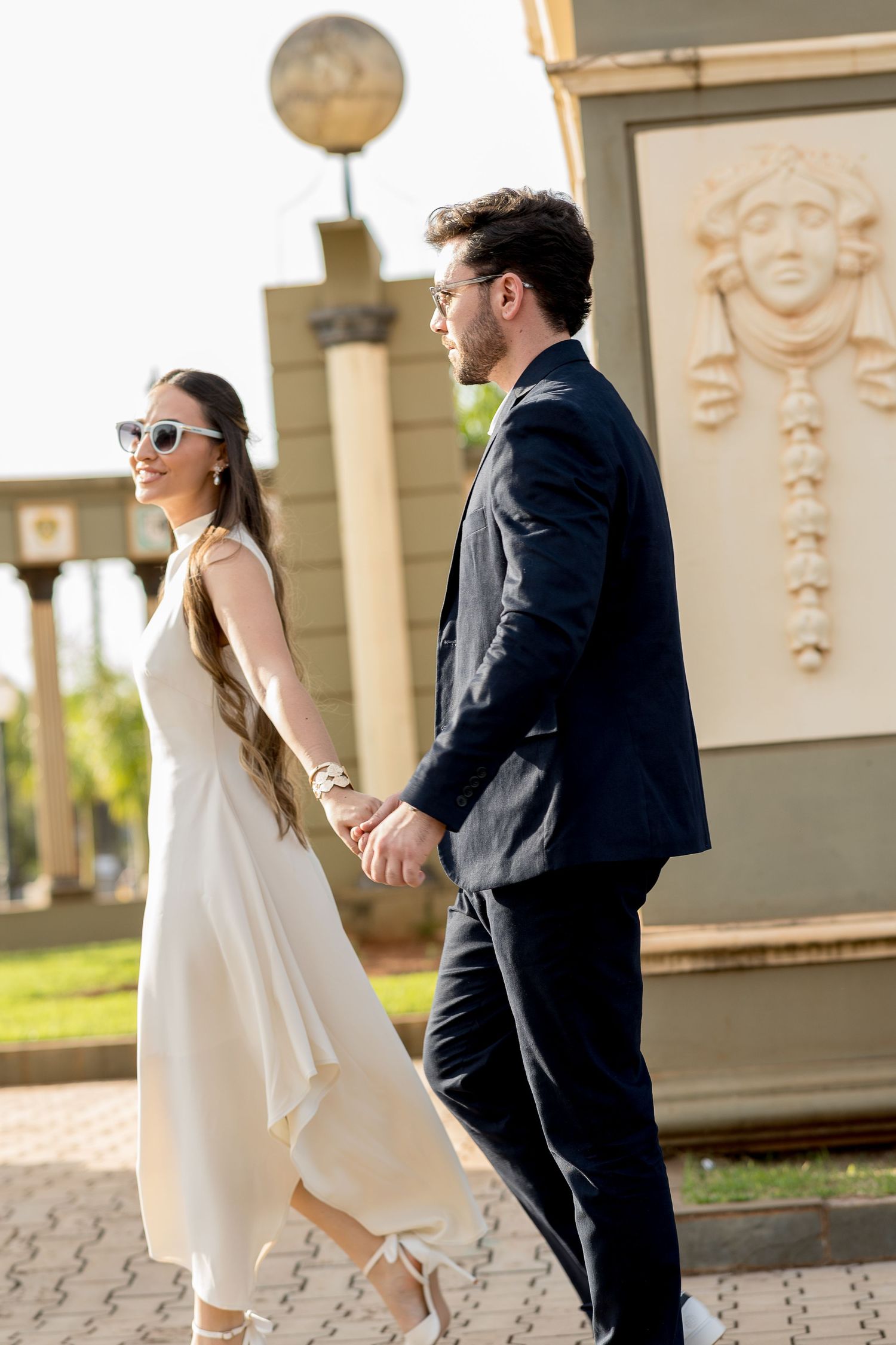 Elegant couple holding hands outdoors, woman in white dress with sunglasses, man in dark suit, near decorative sculpture.