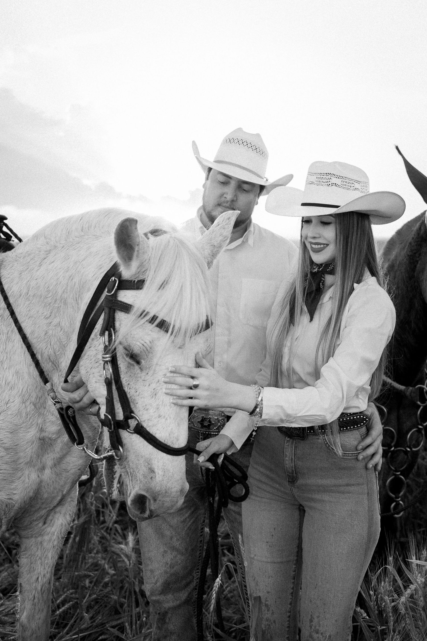 Black and white photo of a couple in cowboy hats petting a white horse outdoors.