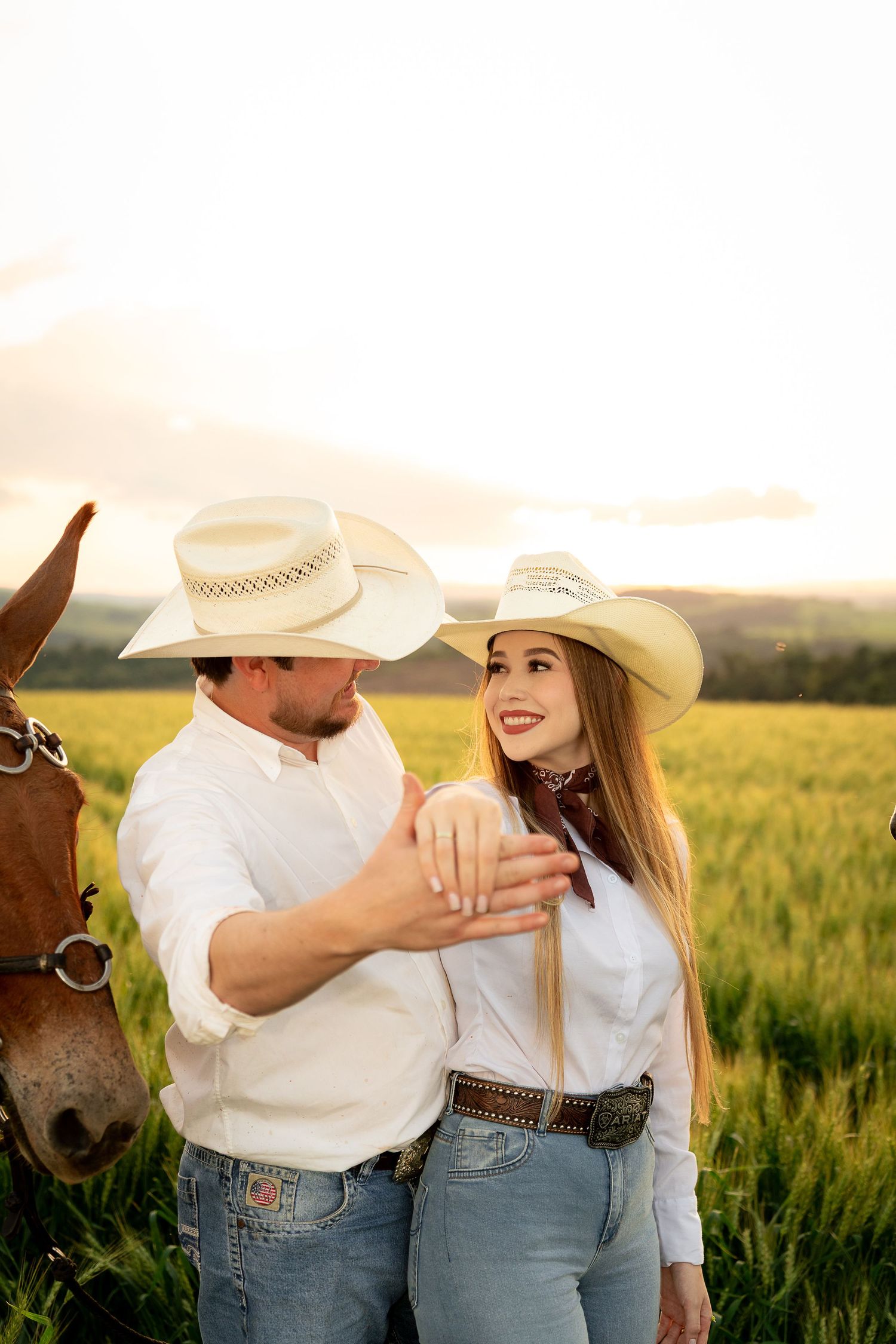 Couple in cowboy hats and white shirts stand together in a golden field at sunset with a horse nearby.
