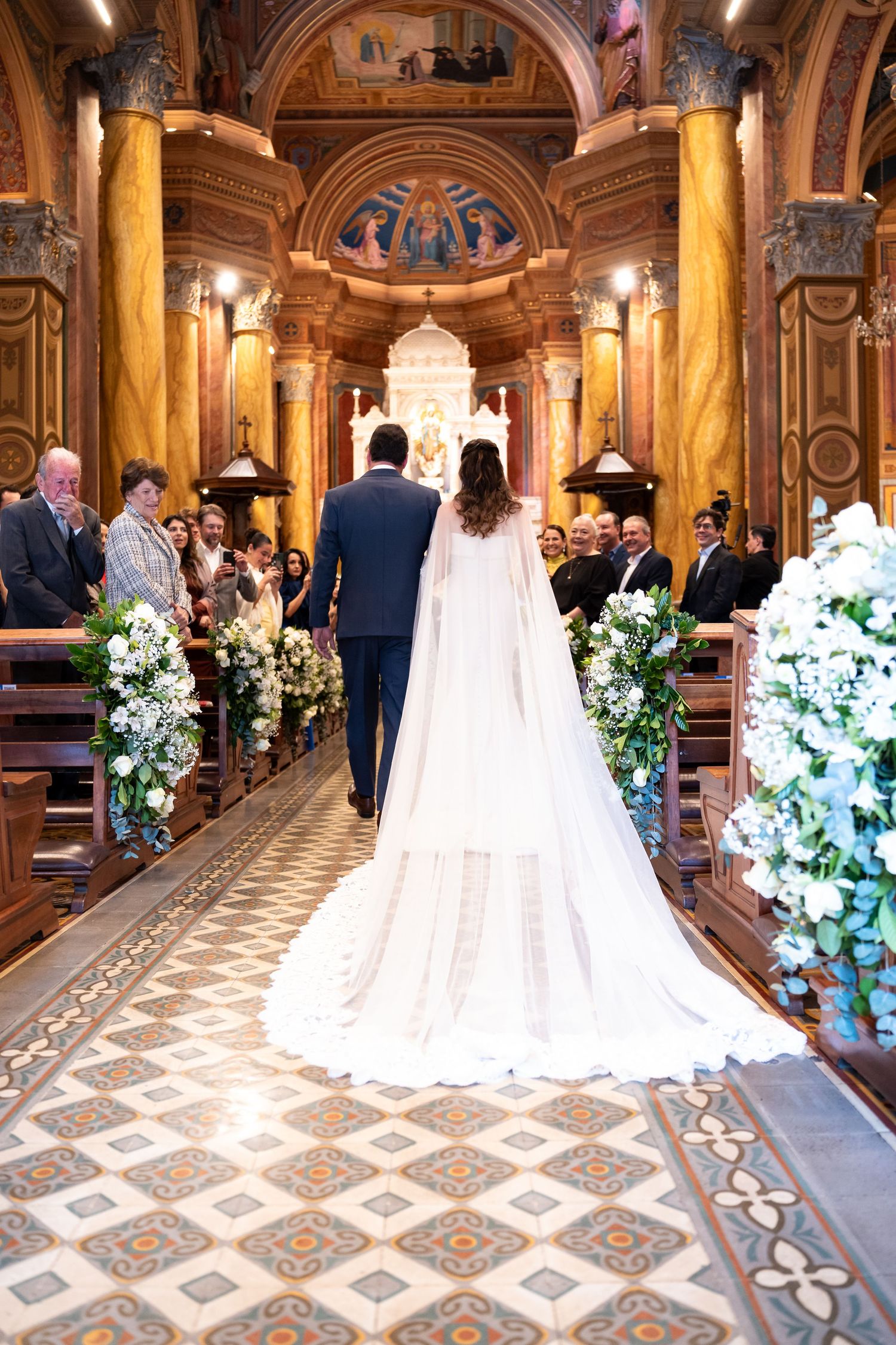 Bride and groom walking down ornate church aisle adorned with white floral arrangements during wedding ceremony.