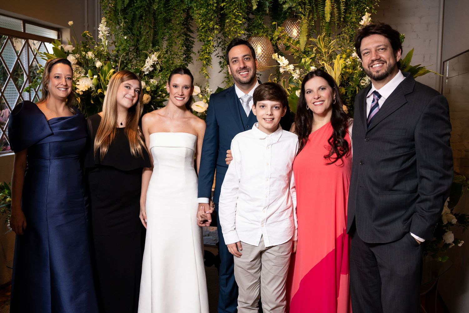 Wedding party posing together in front of a floral backdrop, including bride in white gown and guests in formal attire.