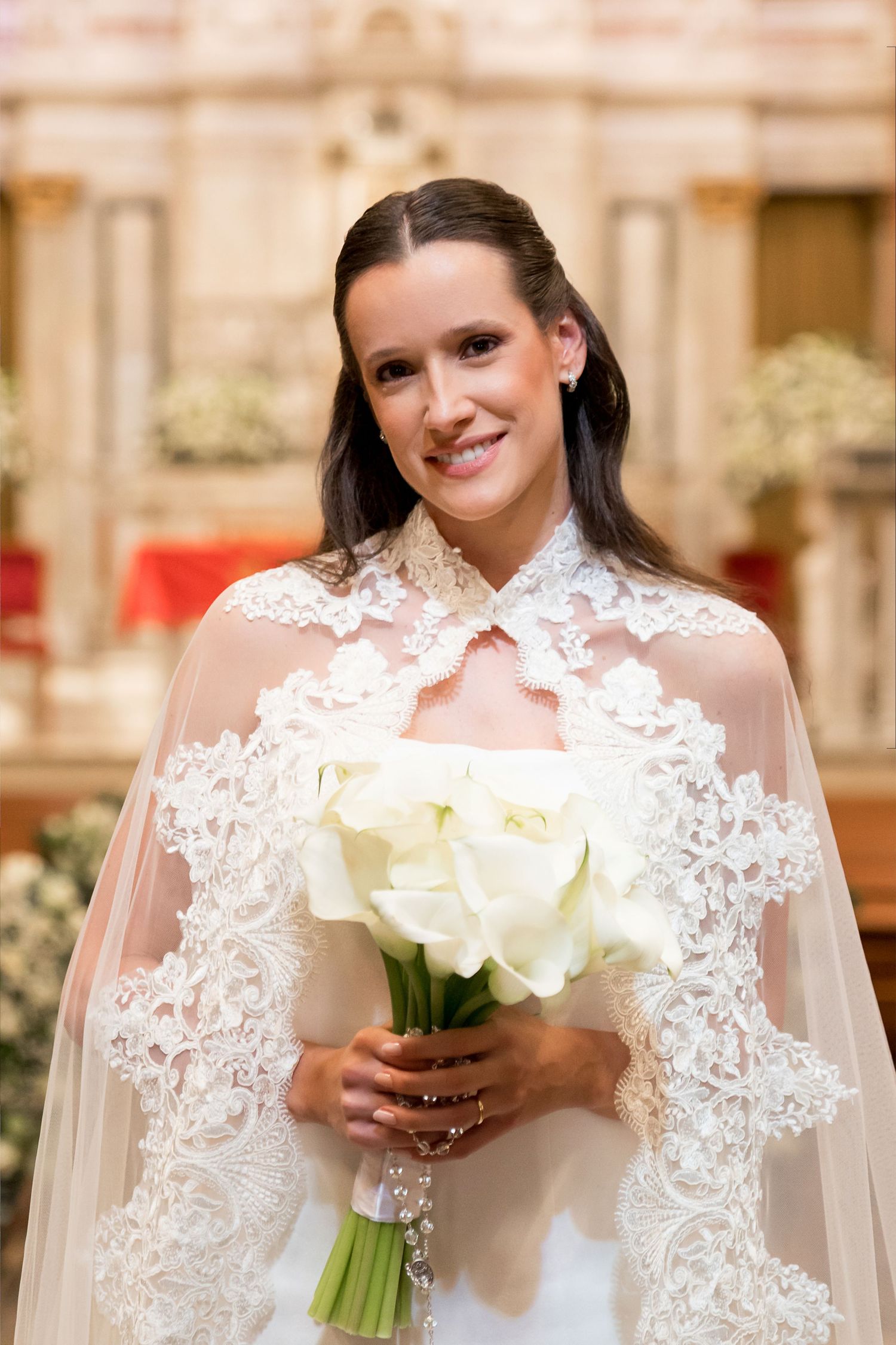 Smiling bride in white lace cape holding elegant calla lily bouquet inside a church.