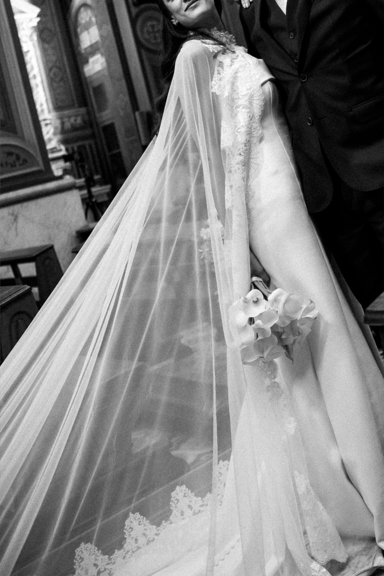 Black and white photo of bride in lace wedding gown with long veil holding bouquet beside groom in church.
