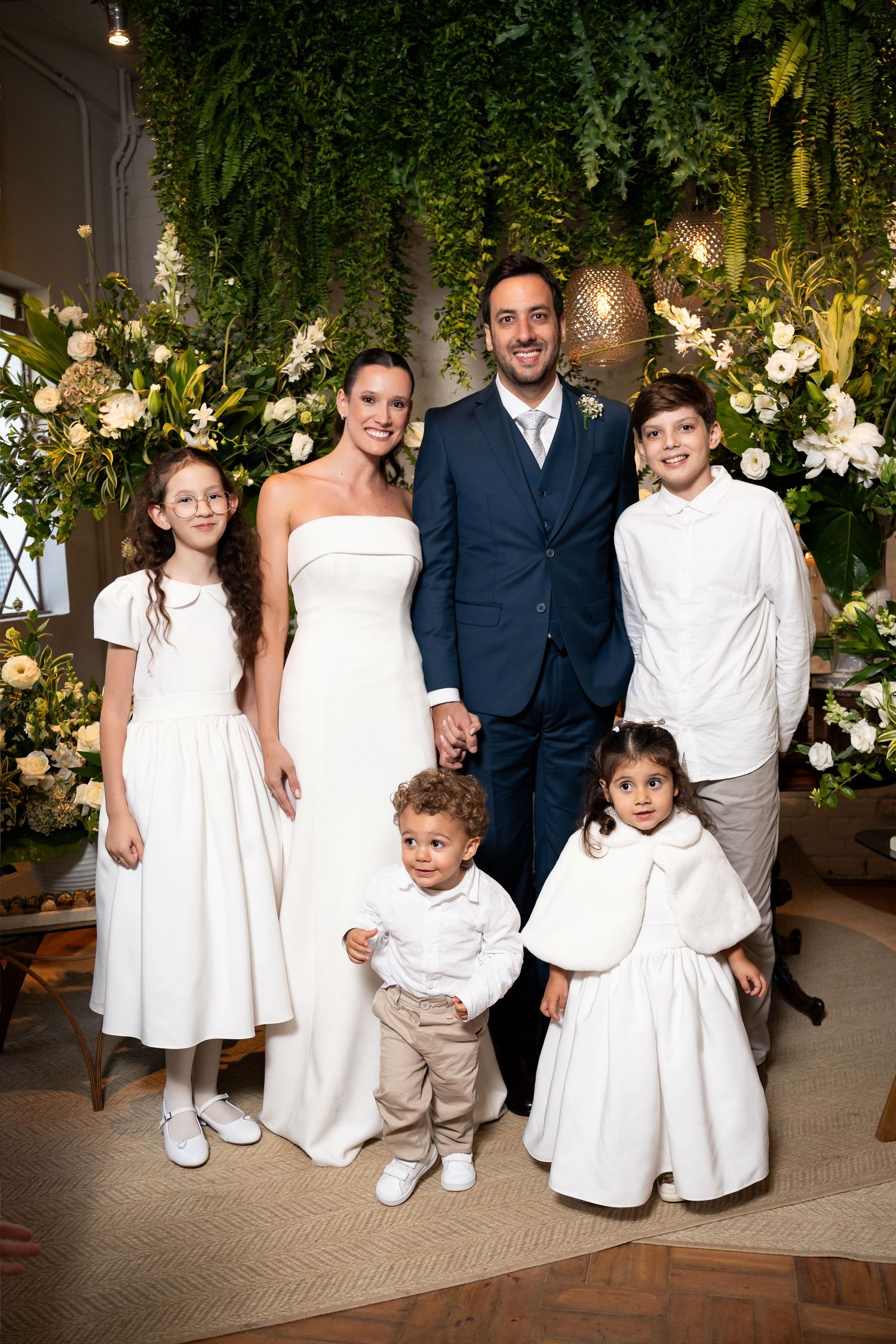Bride and groom pose with four children in white outfits at an elegant wedding ceremony with floral backdrop.