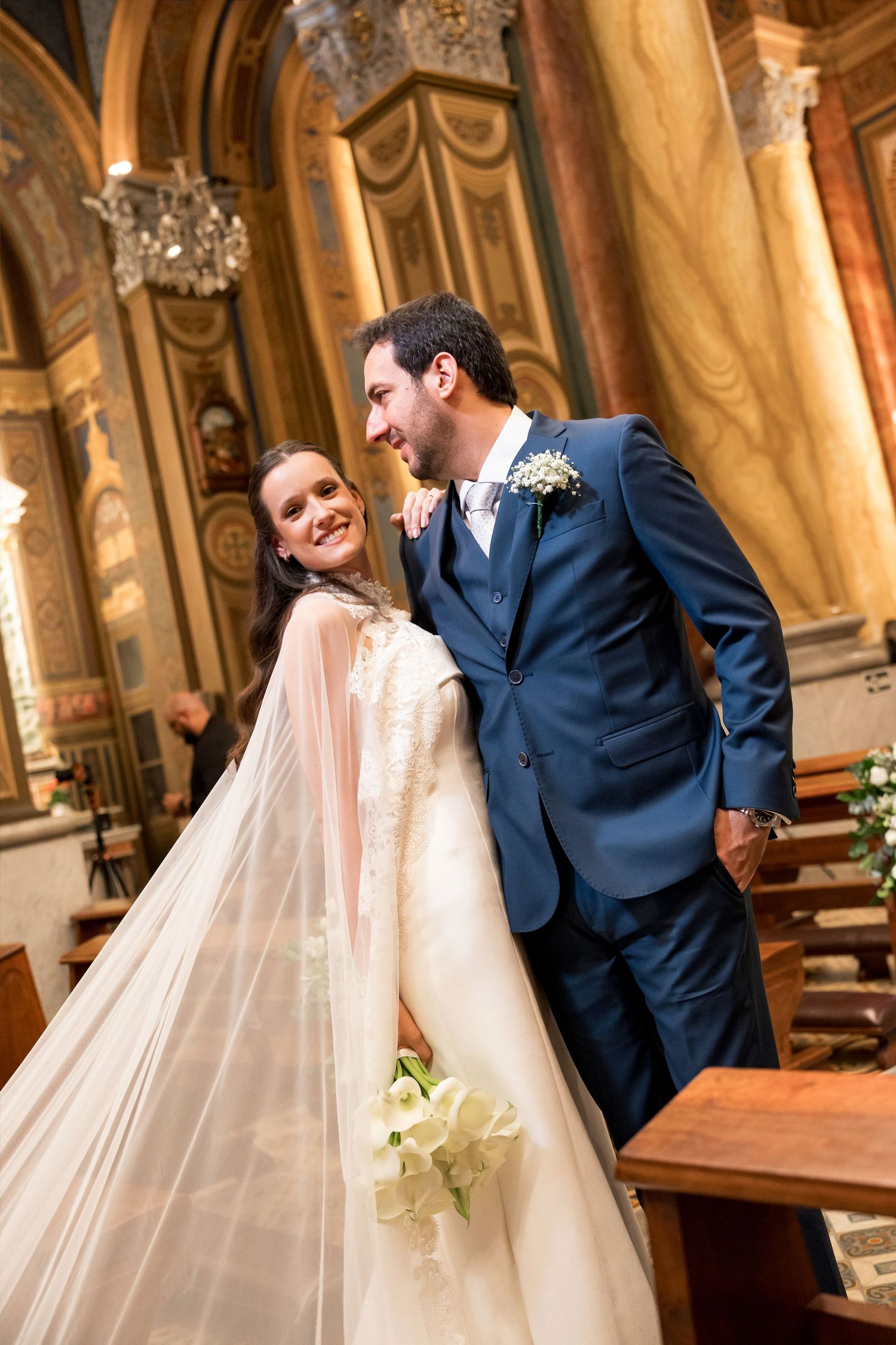 A smiling bride in a white gown with veil and groom in blue suit share a joyful moment inside an ornate church.