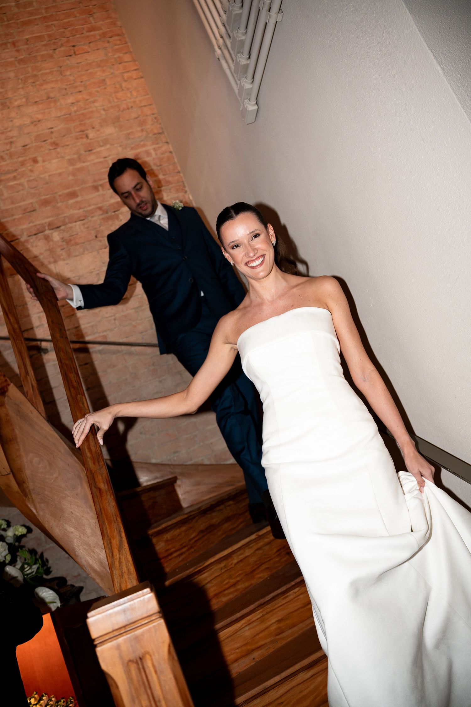 Smiling bride in white strapless gown descends wooden staircase with groom in navy suit behind her.