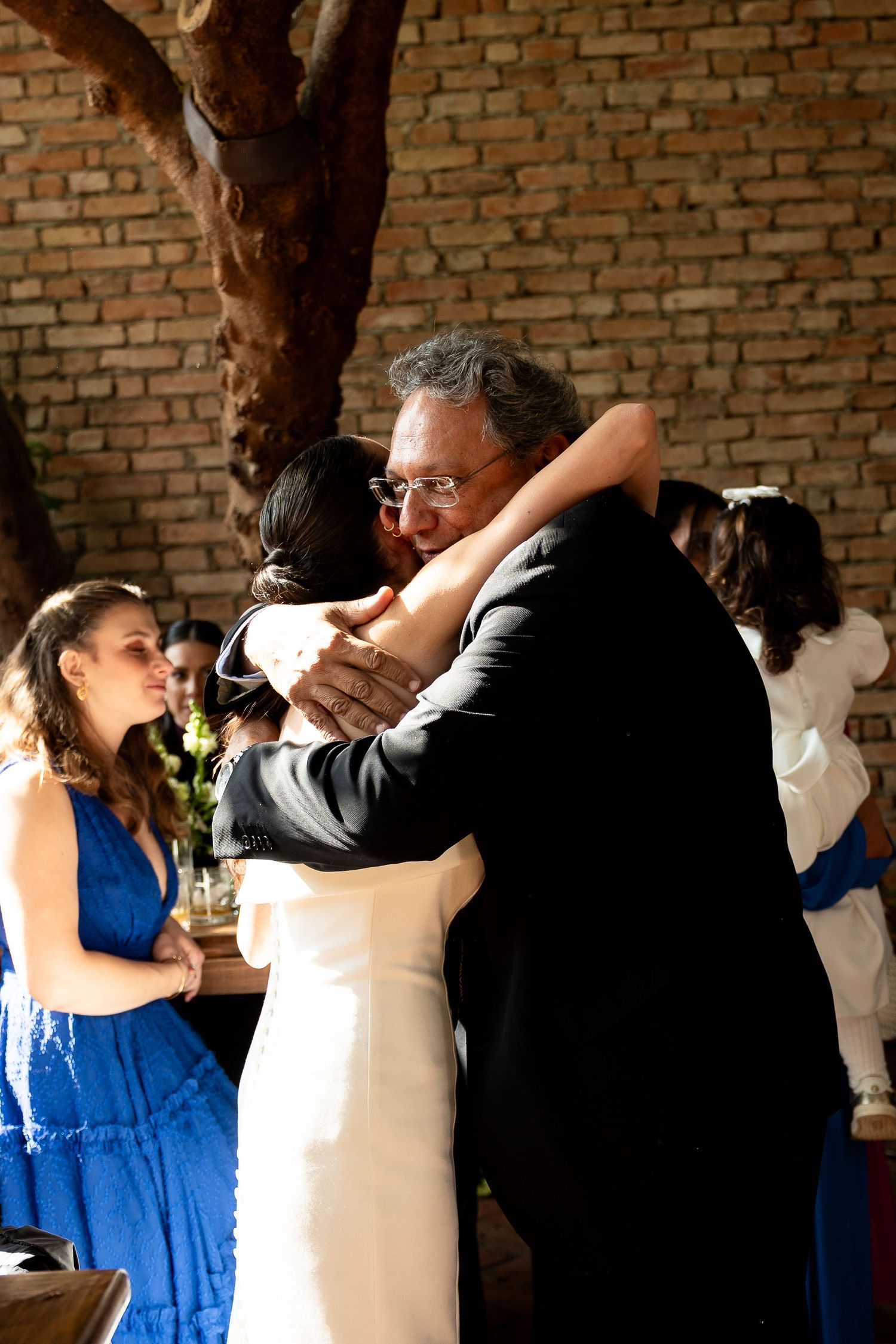 Bride and older man share an emotional hug at wedding reception, with guests in blue dresses watching nearby.