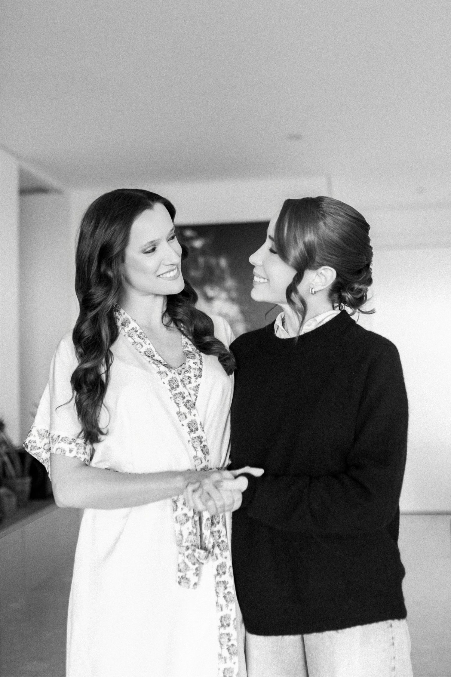 Black and white photo of two women smiling and holding hands, one in a floral robe, sharing a joyful moment.