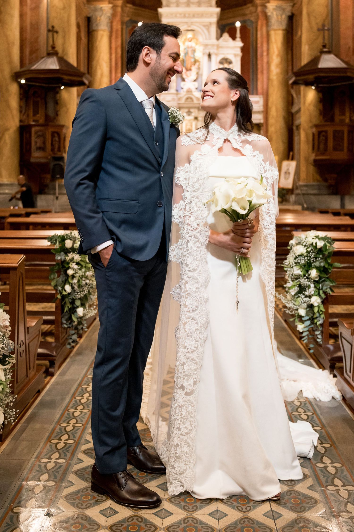 Bride and groom share a romantic moment in a church, bride in lace cape gown holding white roses bouquet.