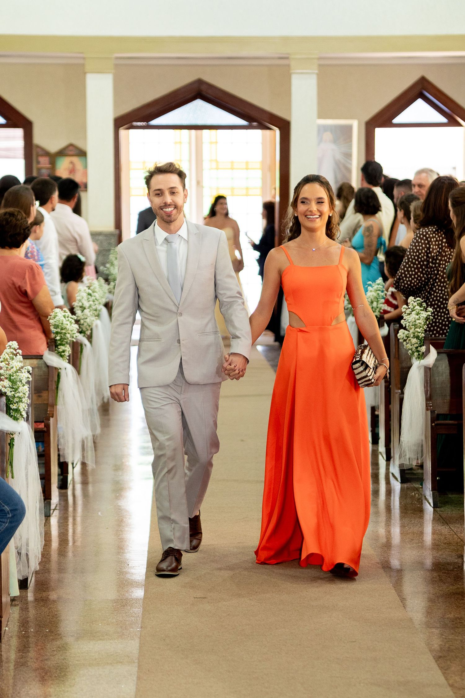 Couple walking down church aisle, man in gray suit, woman in elegant orange gown, holding hands at wedding ceremony.