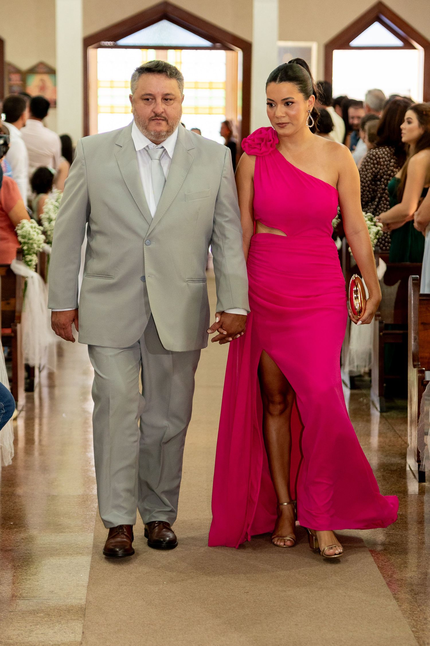 Couple walking down church aisle, man in gray suit, woman in pink one-shoulder gown with slit, wedding ceremony.