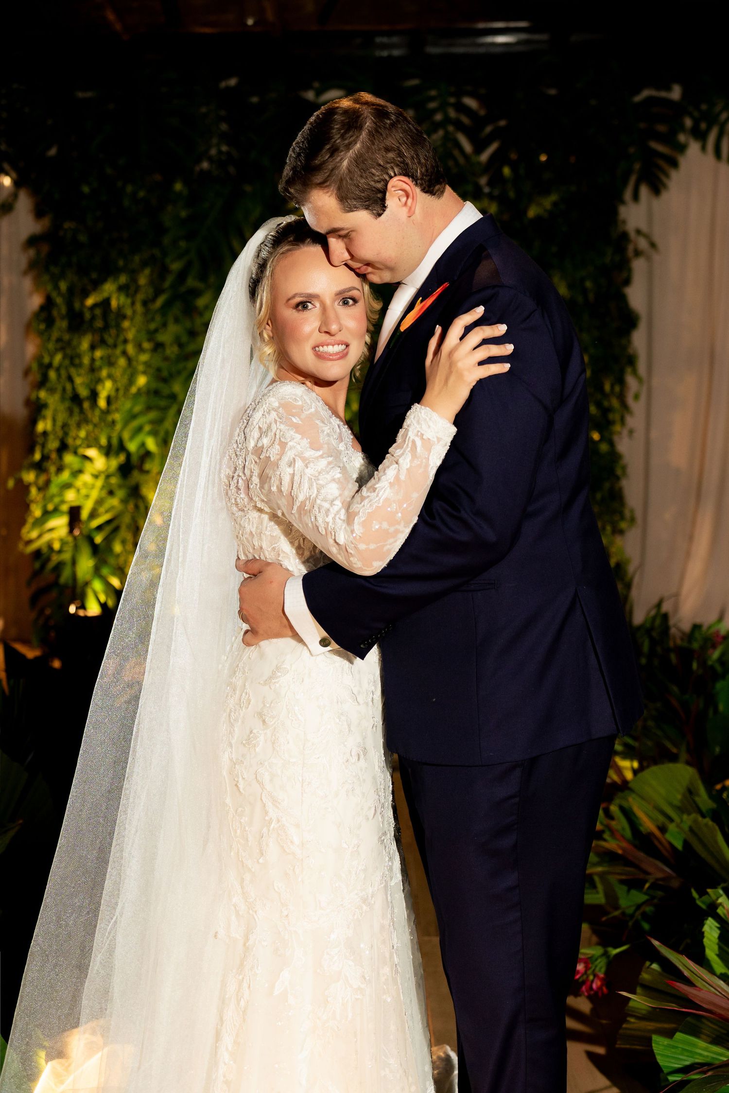 Bride in lace gown and veil embraces groom in navy suit, sharing a romantic moment at their wedding ceremony.