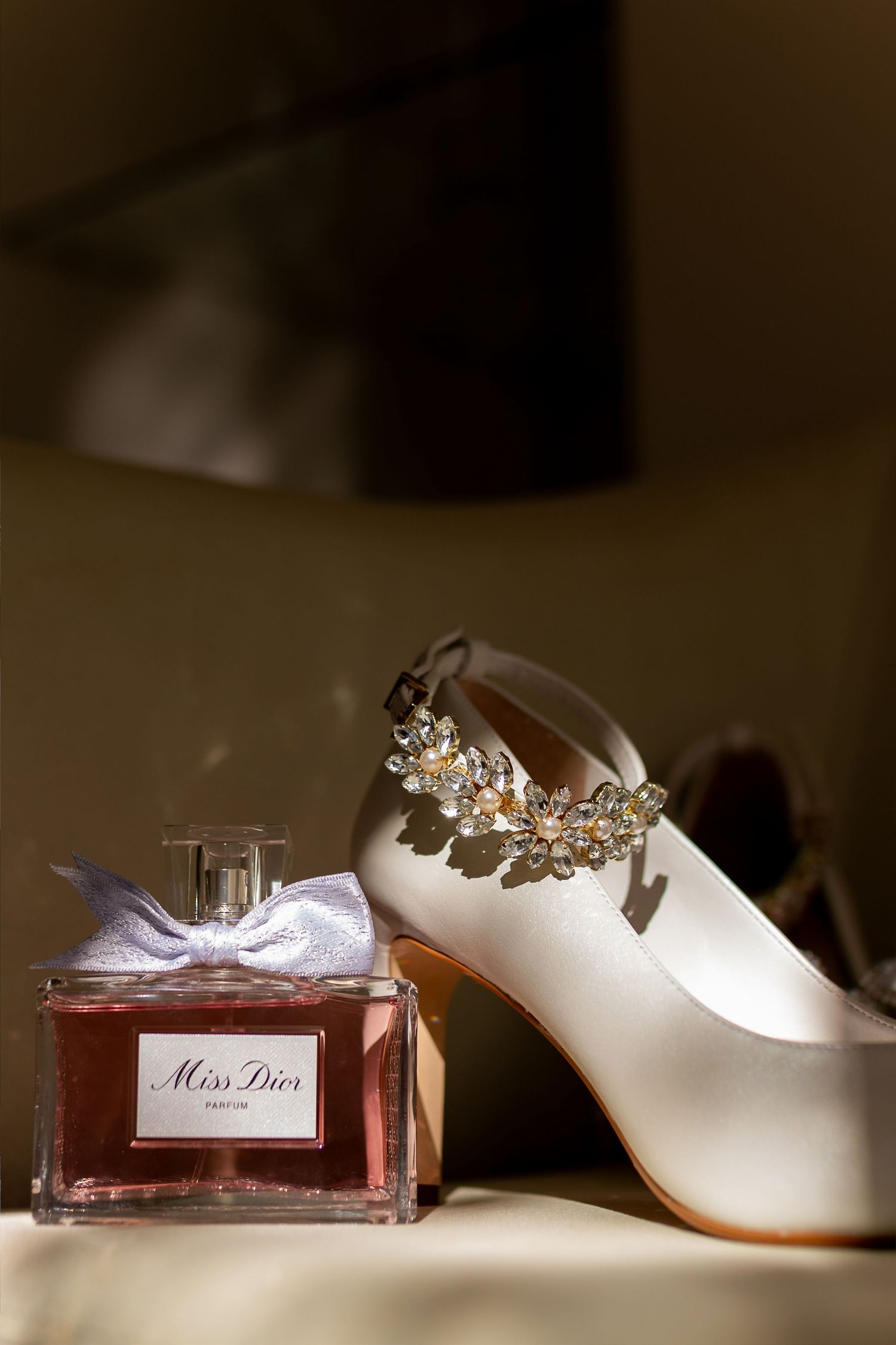 Miss Dior parfum bottle with bow beside white satin bridal heels adorned with crystal floral embellishment.