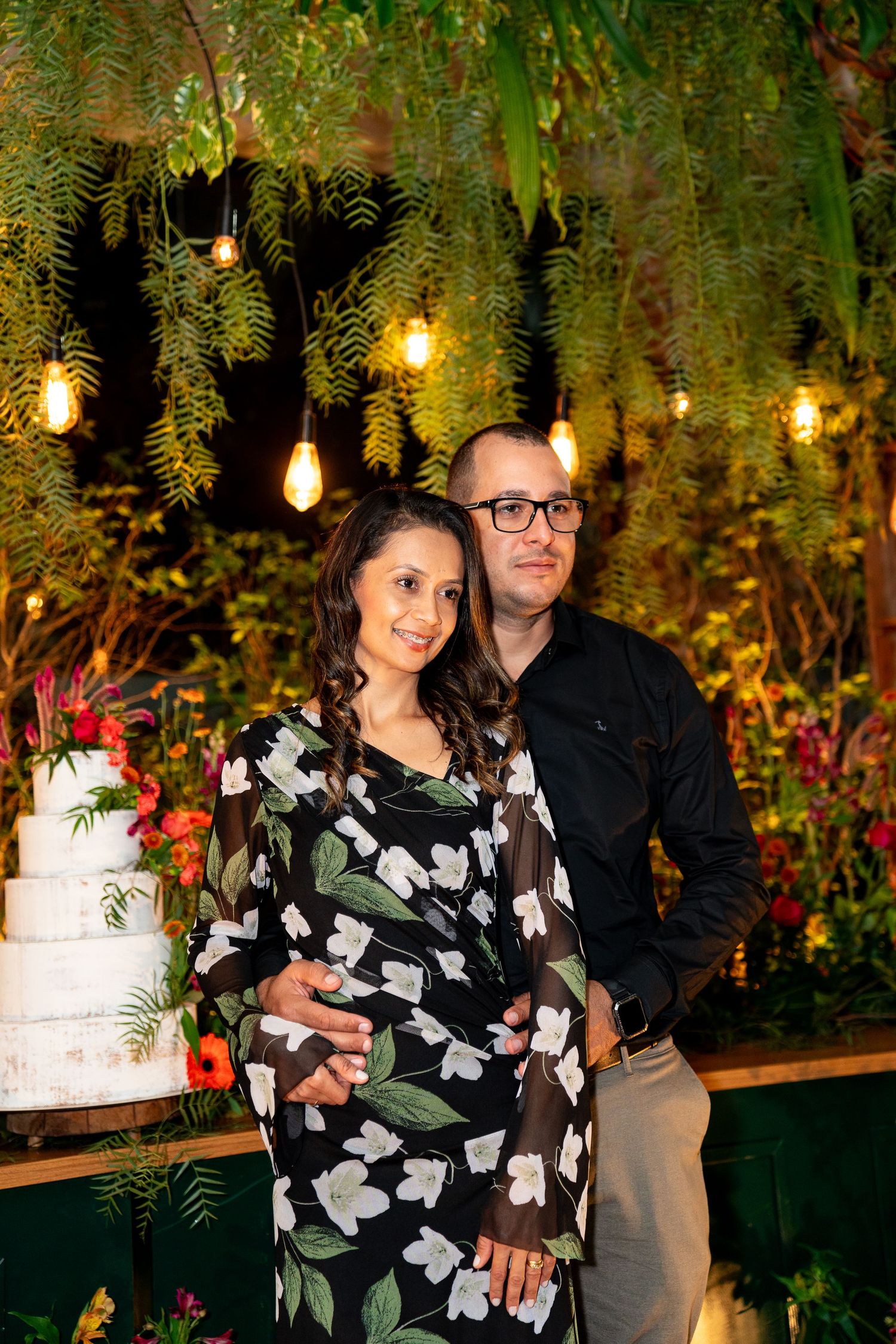 Couple posing together at an elegant event with floral decor, string lights, and a white tiered wedding cake.