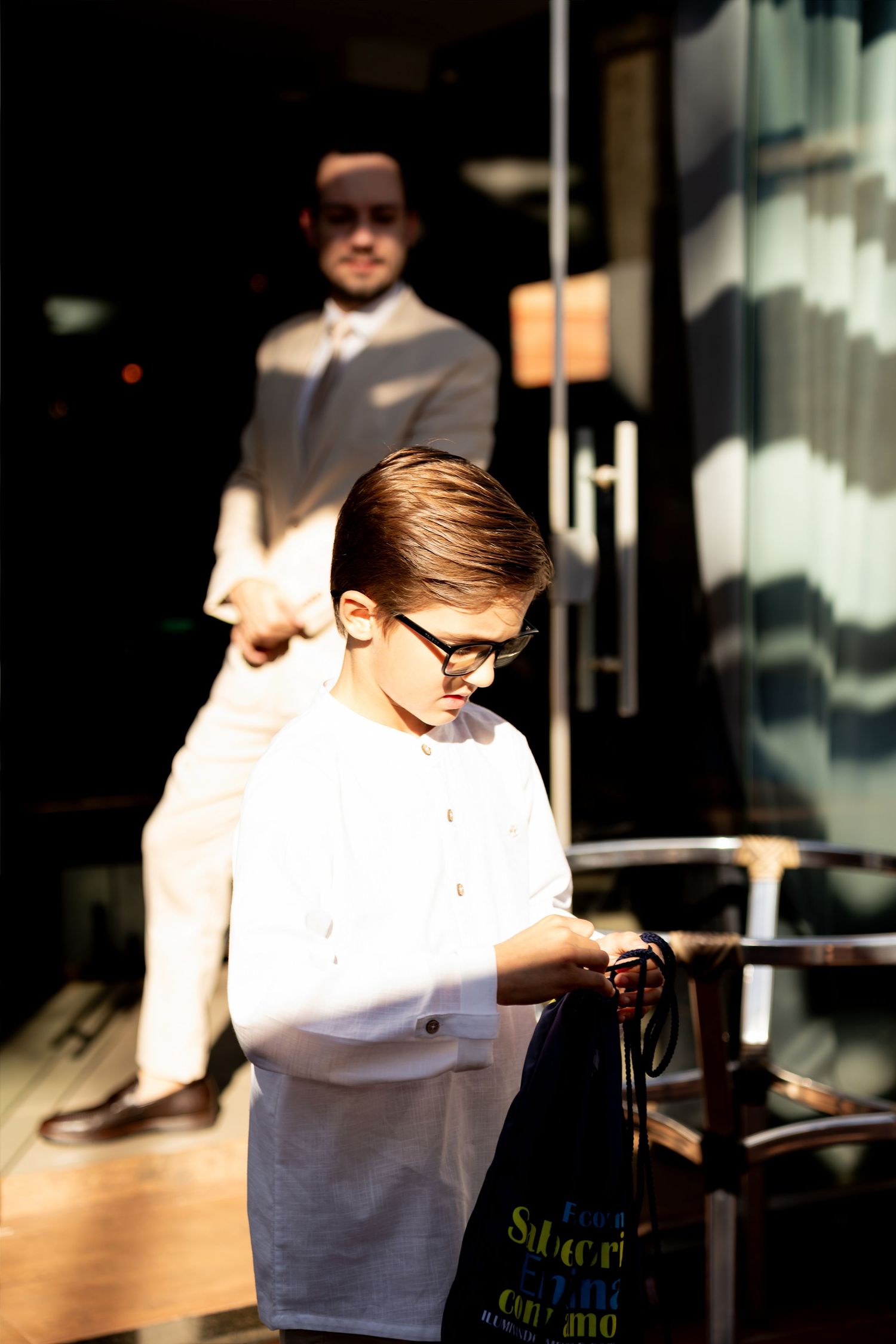 Young boy in white shirt and glasses holding a bag, with a suited man standing in the background near a glass door.