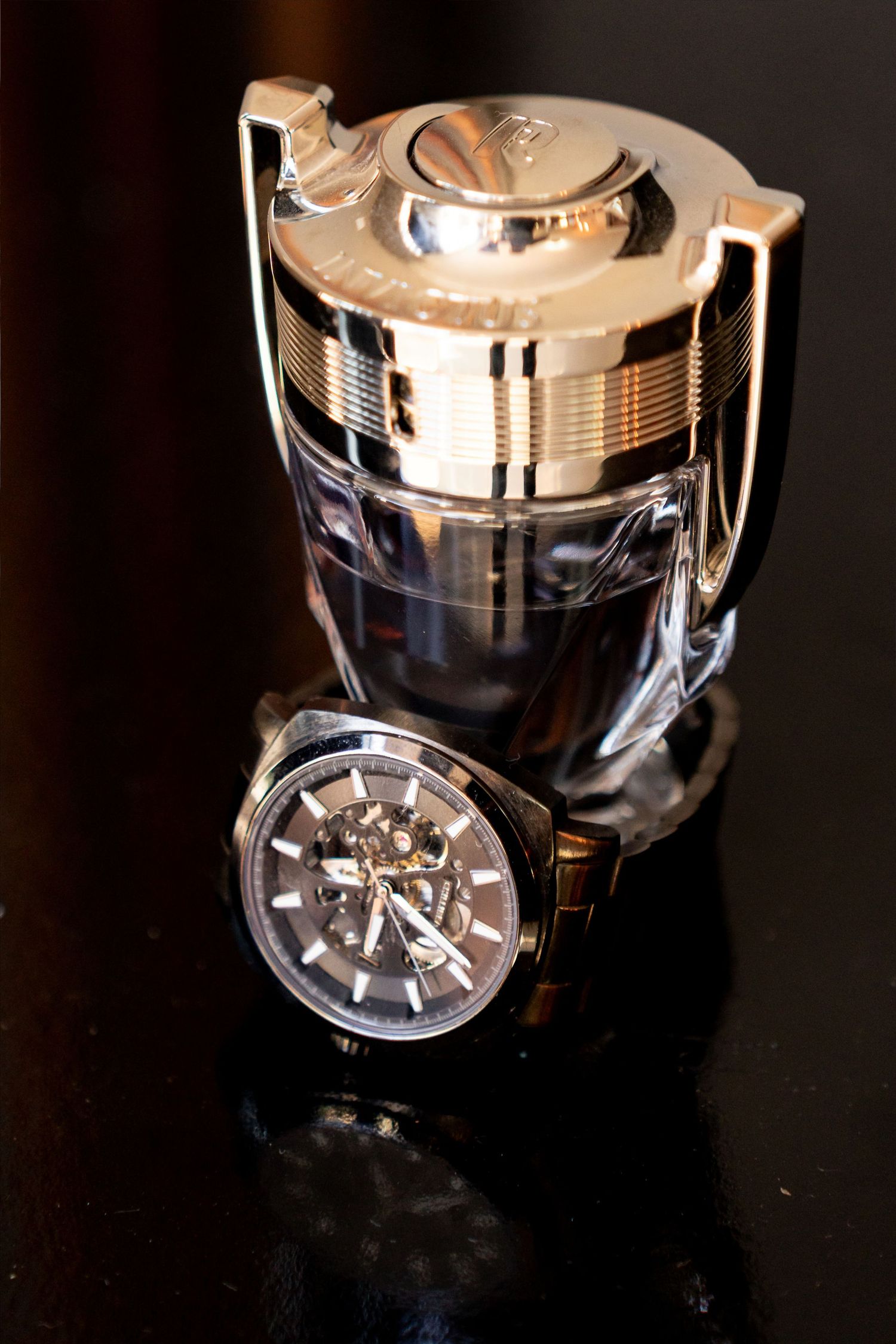 Luxury skeleton wristwatch leaning against a silver trophy-shaped perfume bottle on a dark reflective surface.