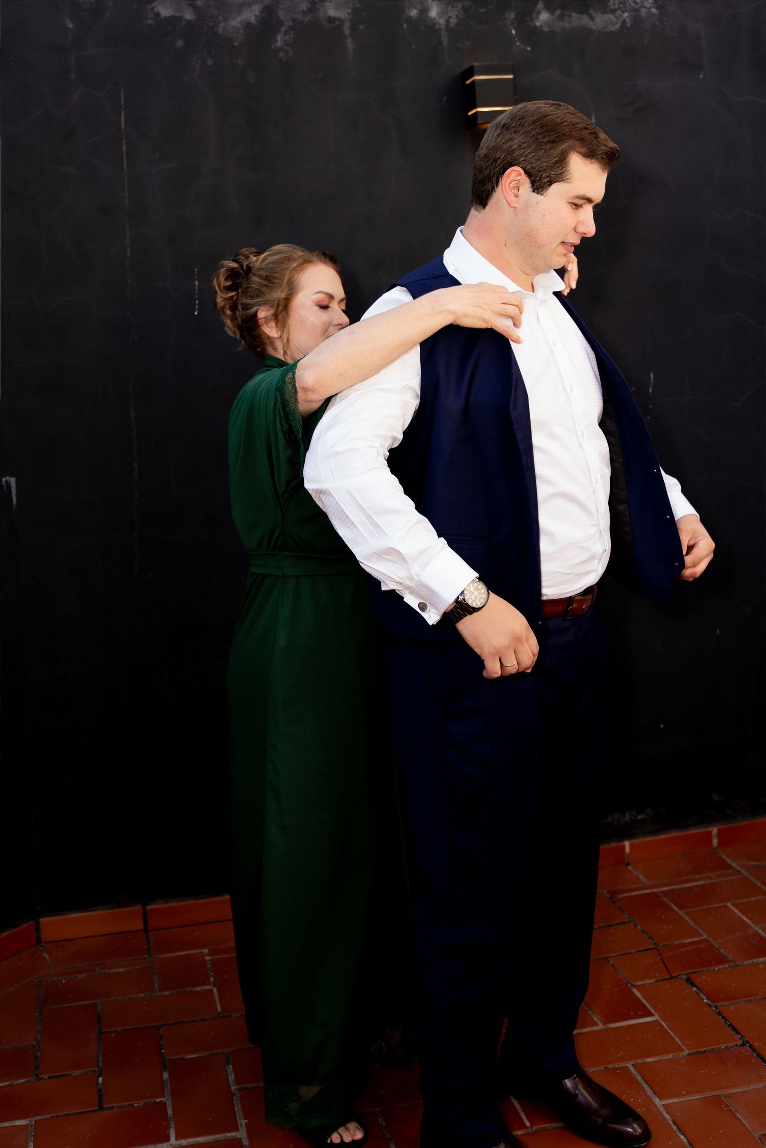 Woman in green dress helping man in navy suit jacket near dark wall on brick floor.