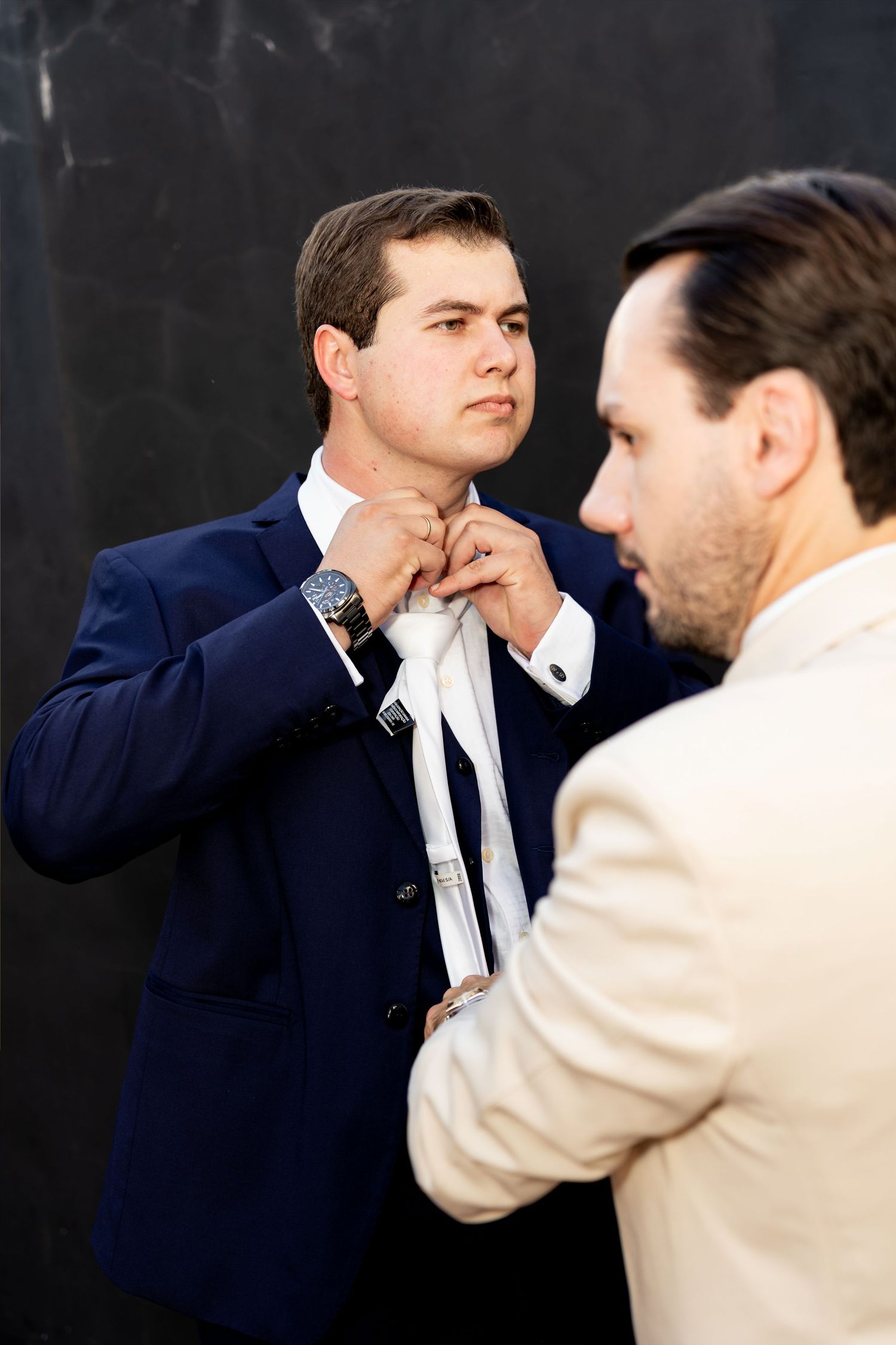 Groomsman in beige suit helping groom in navy suit fix his white tie against dark wall.