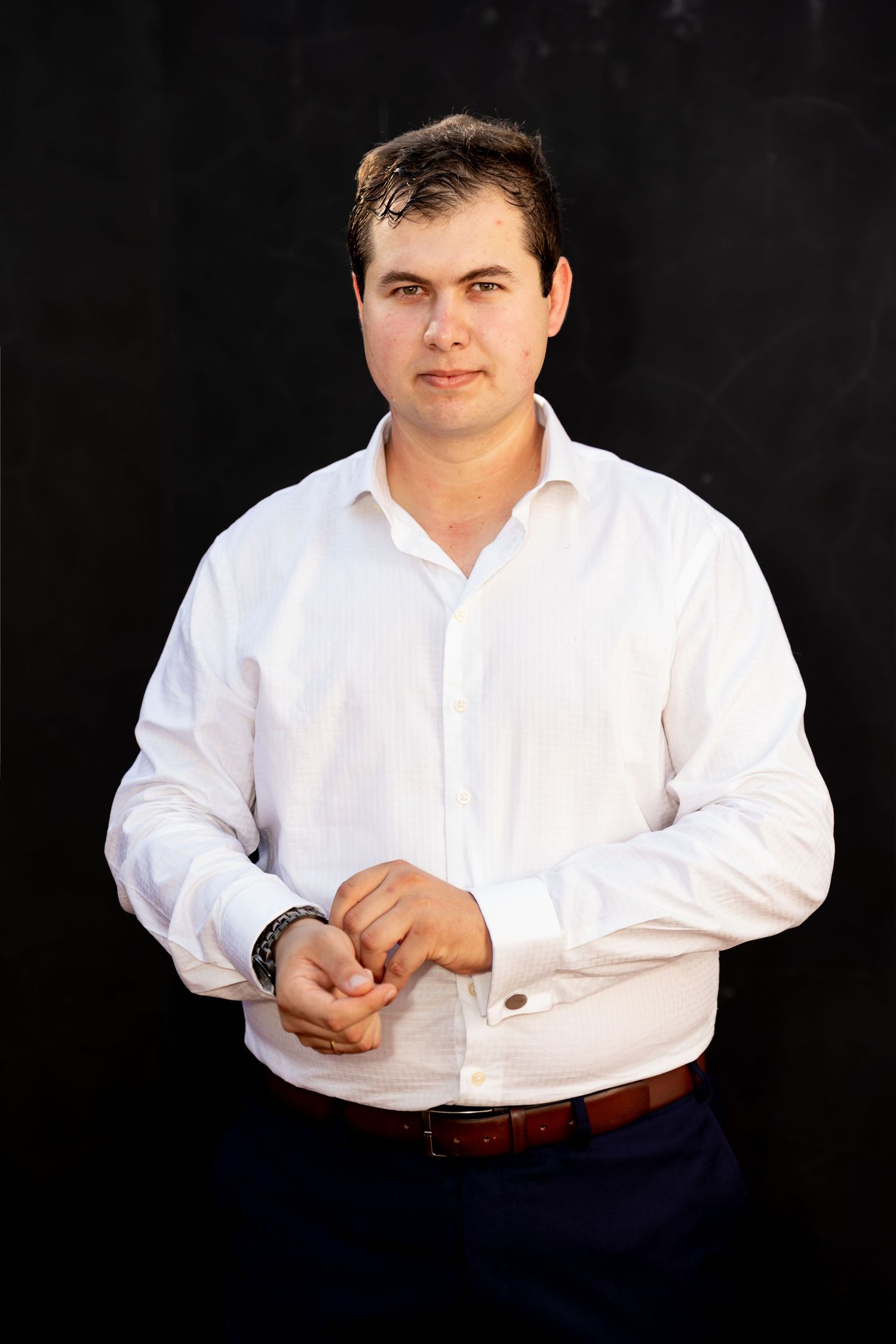 A man in a white dress shirt adjusting his cufflinks against a dark background.