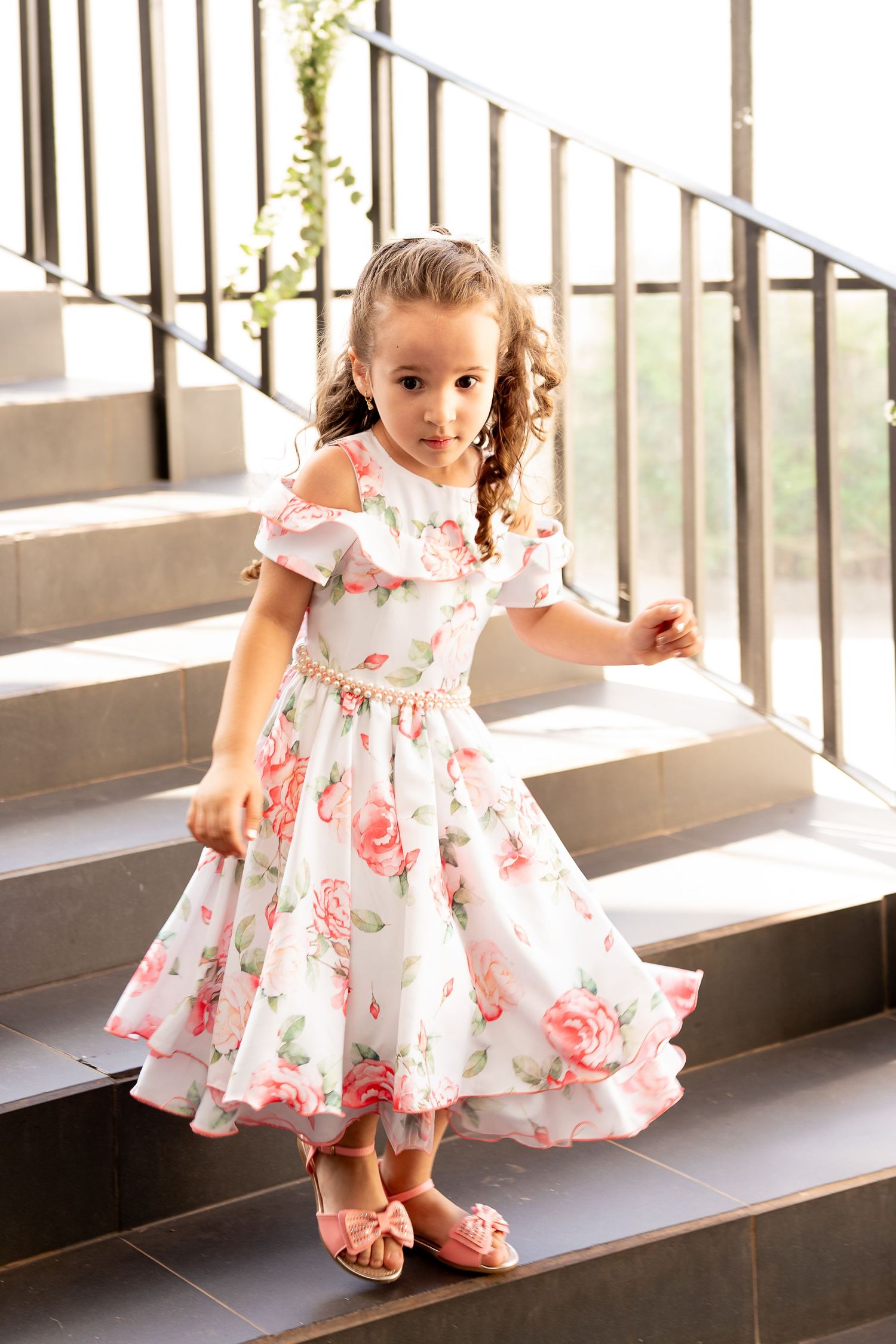 Young girl in white floral dress with pink roses poses gracefully on stone stairs in natural light.