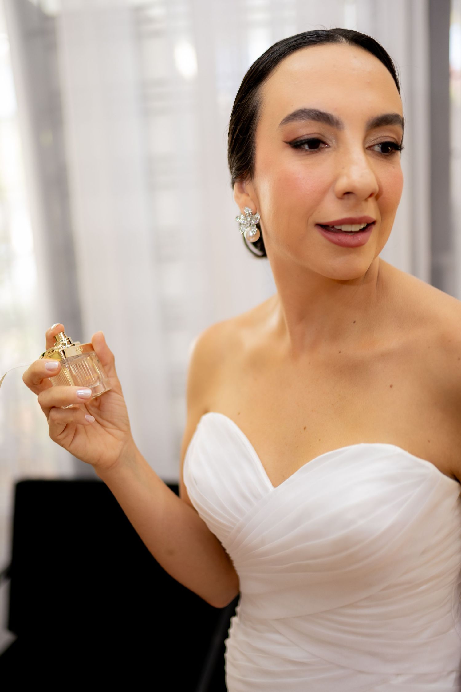 A bride in a white strapless gown holds a perfume bottle, wearing elegant earrings, preparing for her wedding day.