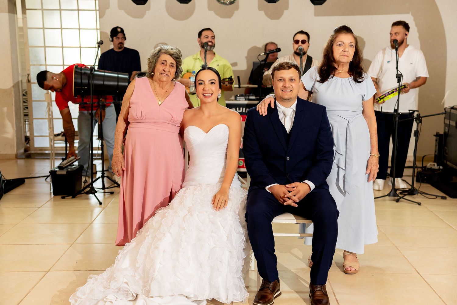 Bride in white gown poses with family and live band at elegant wedding reception venue.